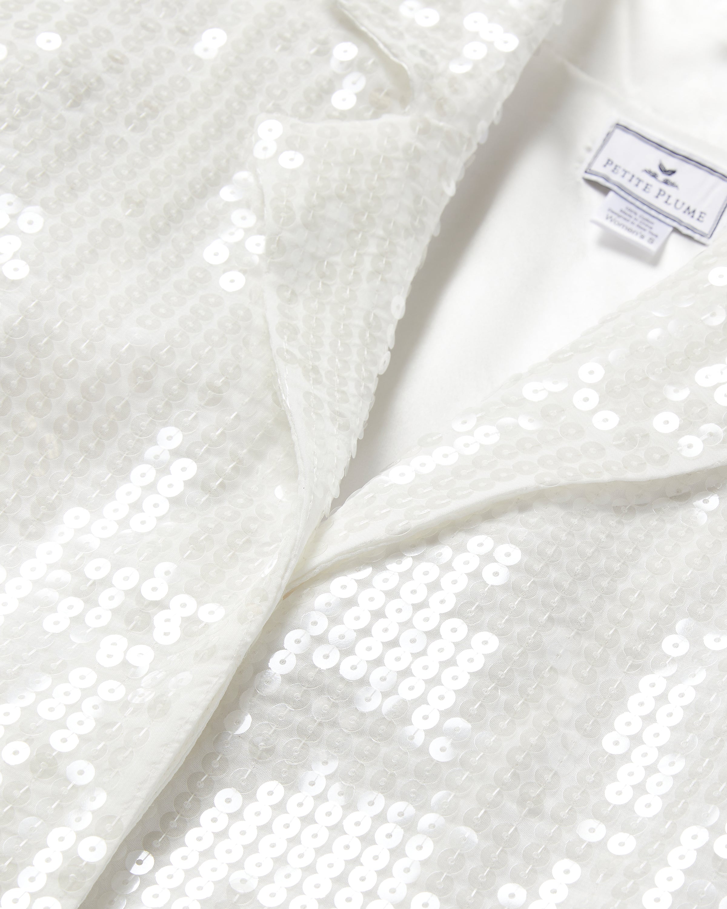 Close-up of Womens Sequin Pajama Set in White by Petite Plume. The sequined fabric has a subtle shine and uniform texture, creating a reflective surface akin to a classic pajama silhouette. A Petite Plume label is visible, emphasizing luxury sleepwear design.