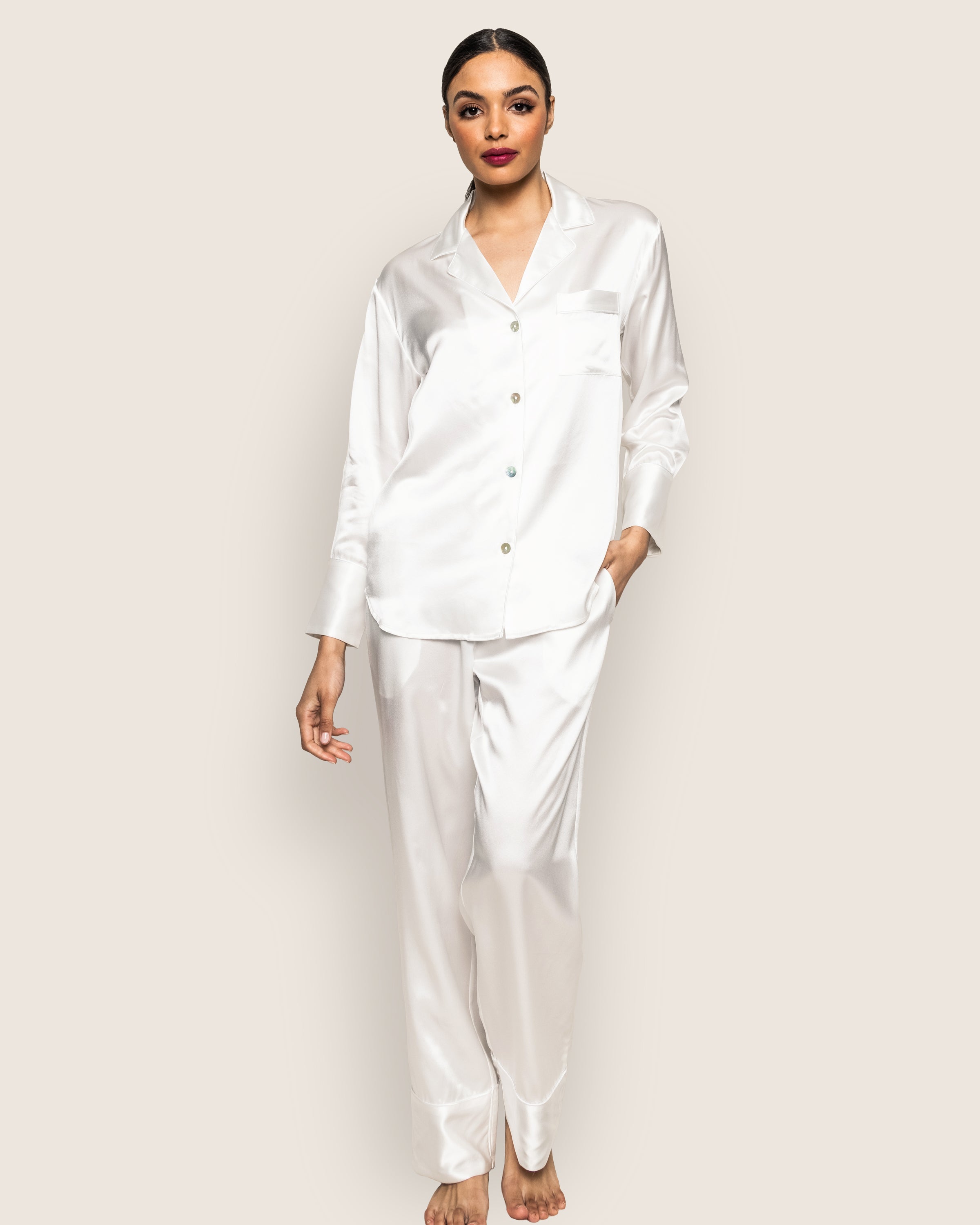 Against a plain background, the individual stands barefoot in a Womens Silk Pajama Set in White by Petite Plume. The elegant set features a button-up shirt with a pocket and matching pants. With hair tied back, they gaze forward with relaxed poise.