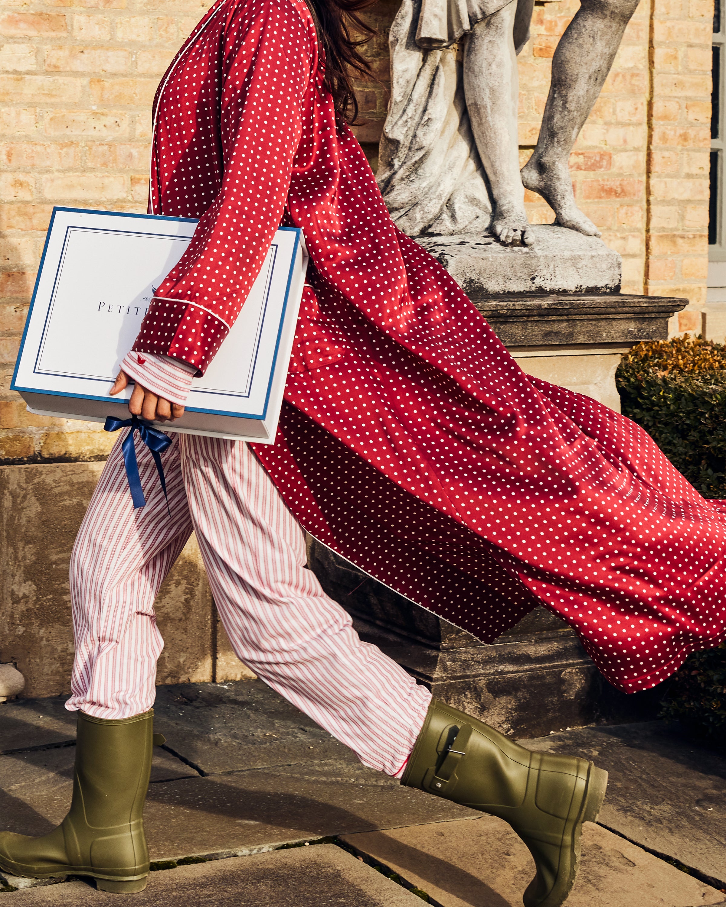 A person in a flowing Womens Silk Long Robe in Bordeaux Polka Dot by Petite Plume walks with two books, wearing striped pants and green boots that echo luxury sleepwear. Stone statues and a brick wall enhance their artful, tranquil stride.