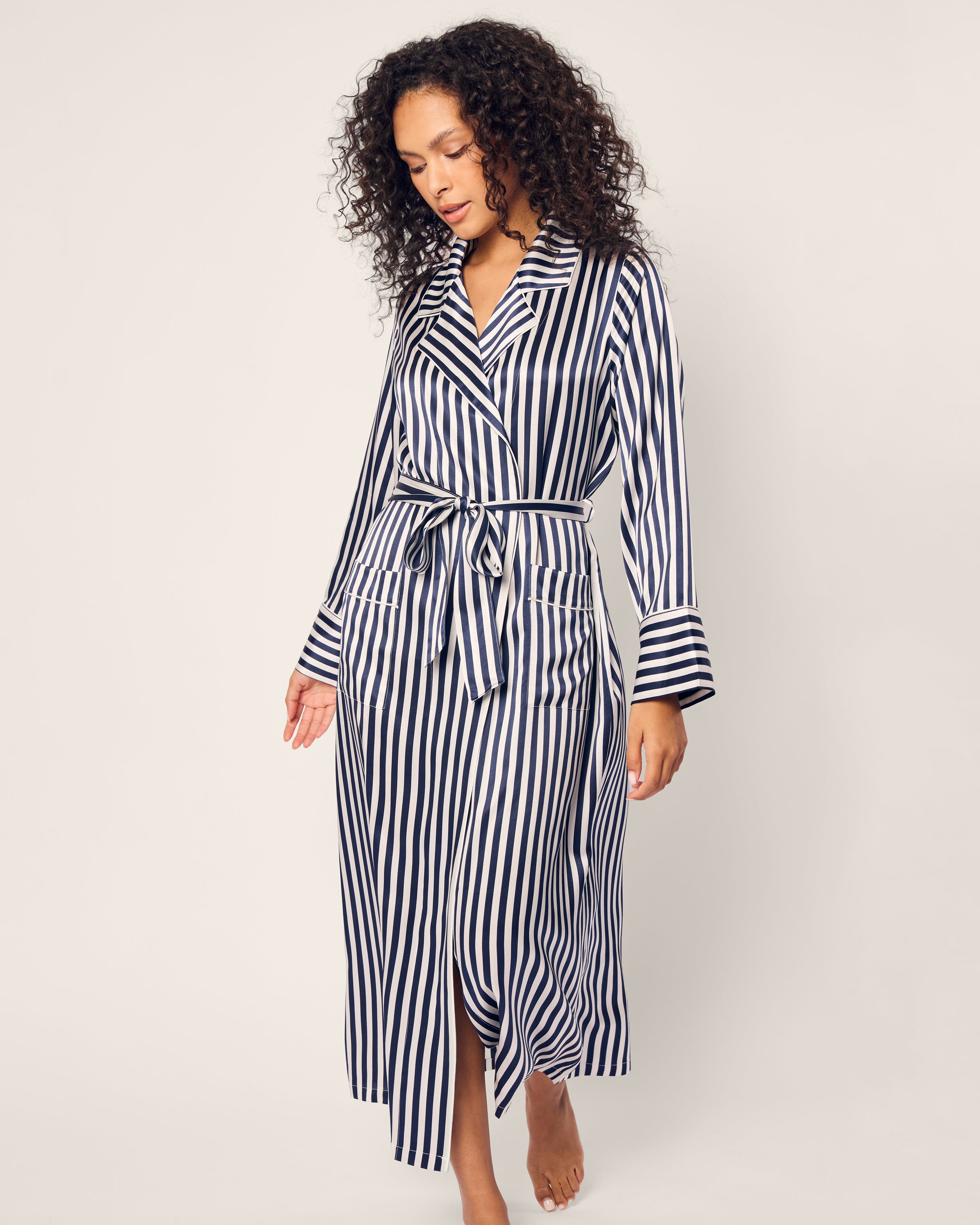 A woman with curly hair wears the Petite Plume Womens Silk Robe in Navy Bengal Stripe, complete with a tie belt and two front pockets. She stands barefoot against a plain background, exuding relaxed elegance.