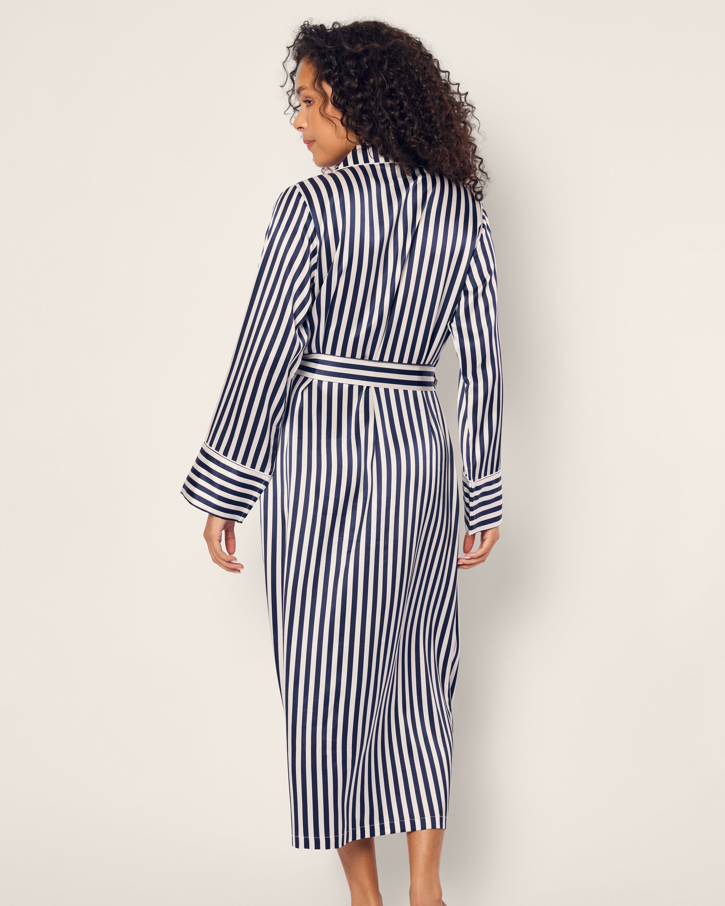 A person with curly hair is wearing Petite Plumes Womens Silk Robe in Navy Bengal Stripe, accented by a belt. The luxurious sleepwear ensemble adds sophistication as they stand against a plain background, facing slightly away from the camera.