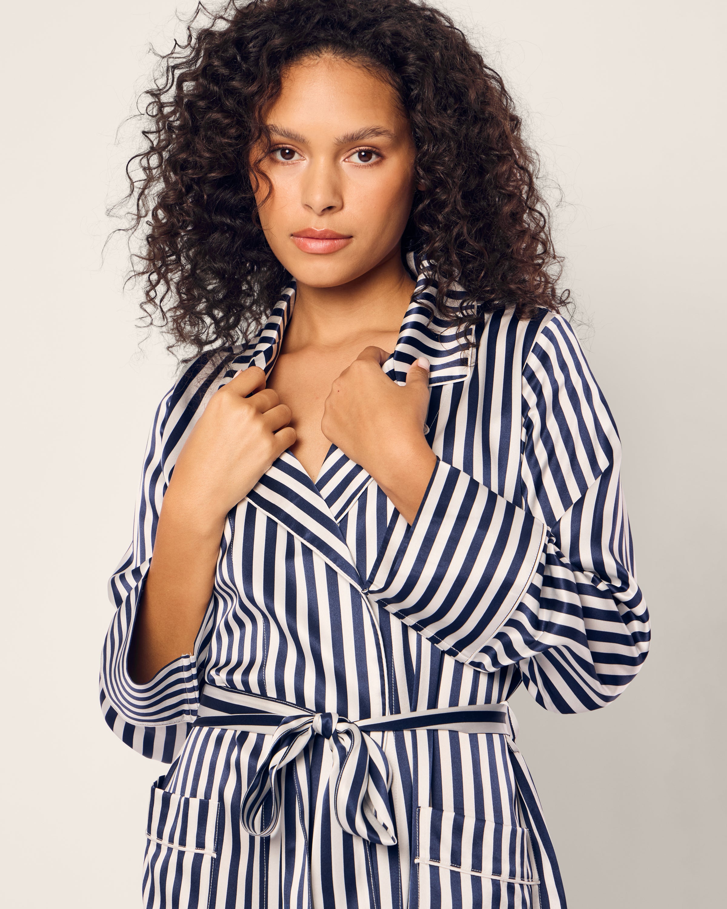 A person with curly hair stands against a plain background, wearing Petite Plumes Womens Silk Robe in Navy Bengal Stripe. They hold the collar with both hands, exuding elegance and comfort while looking directly at the camera.