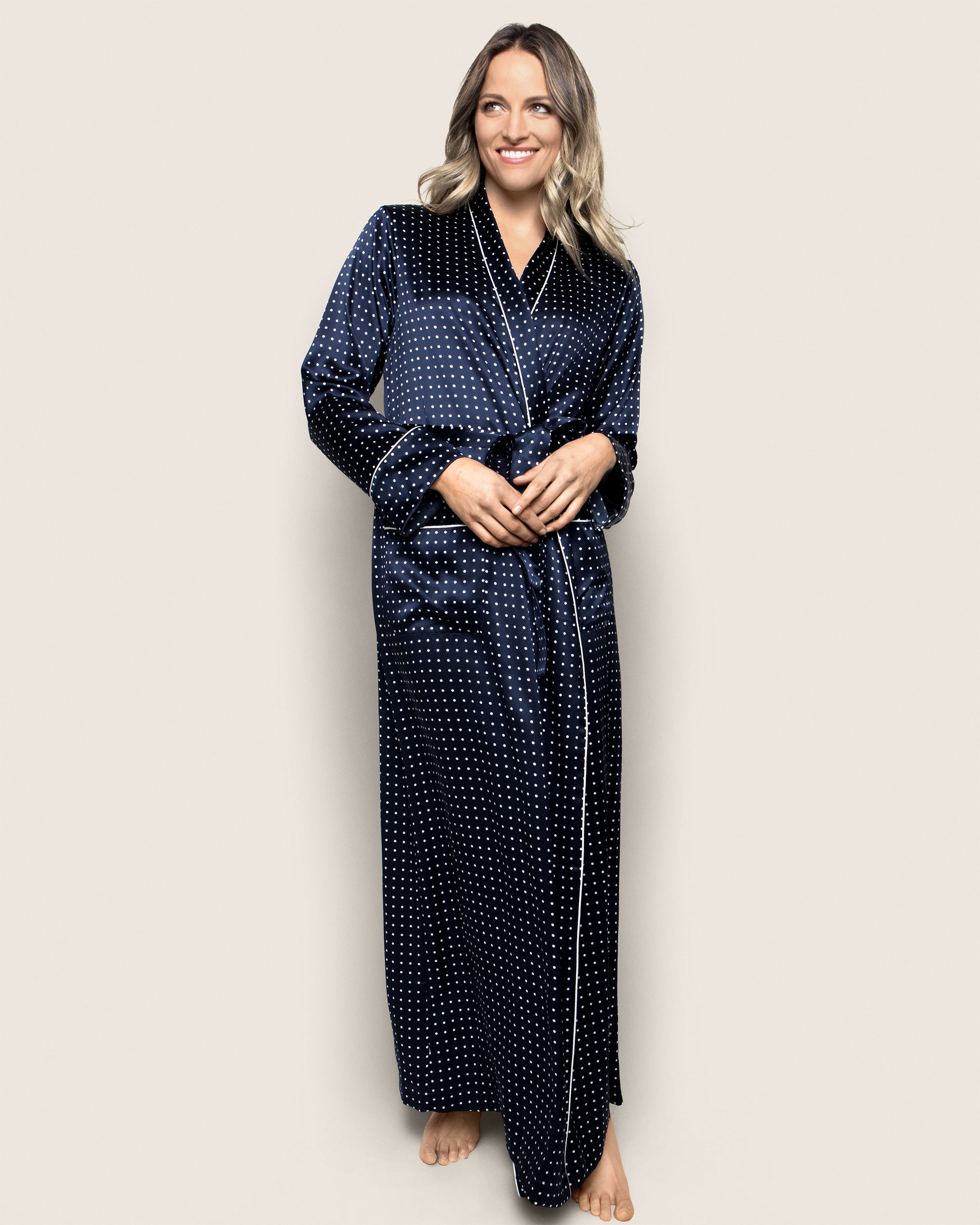 A person stands barefoot, smiling in Petite Plumes Womens Silk Long Robe in Navy Polka Dot. The background is plain beige.