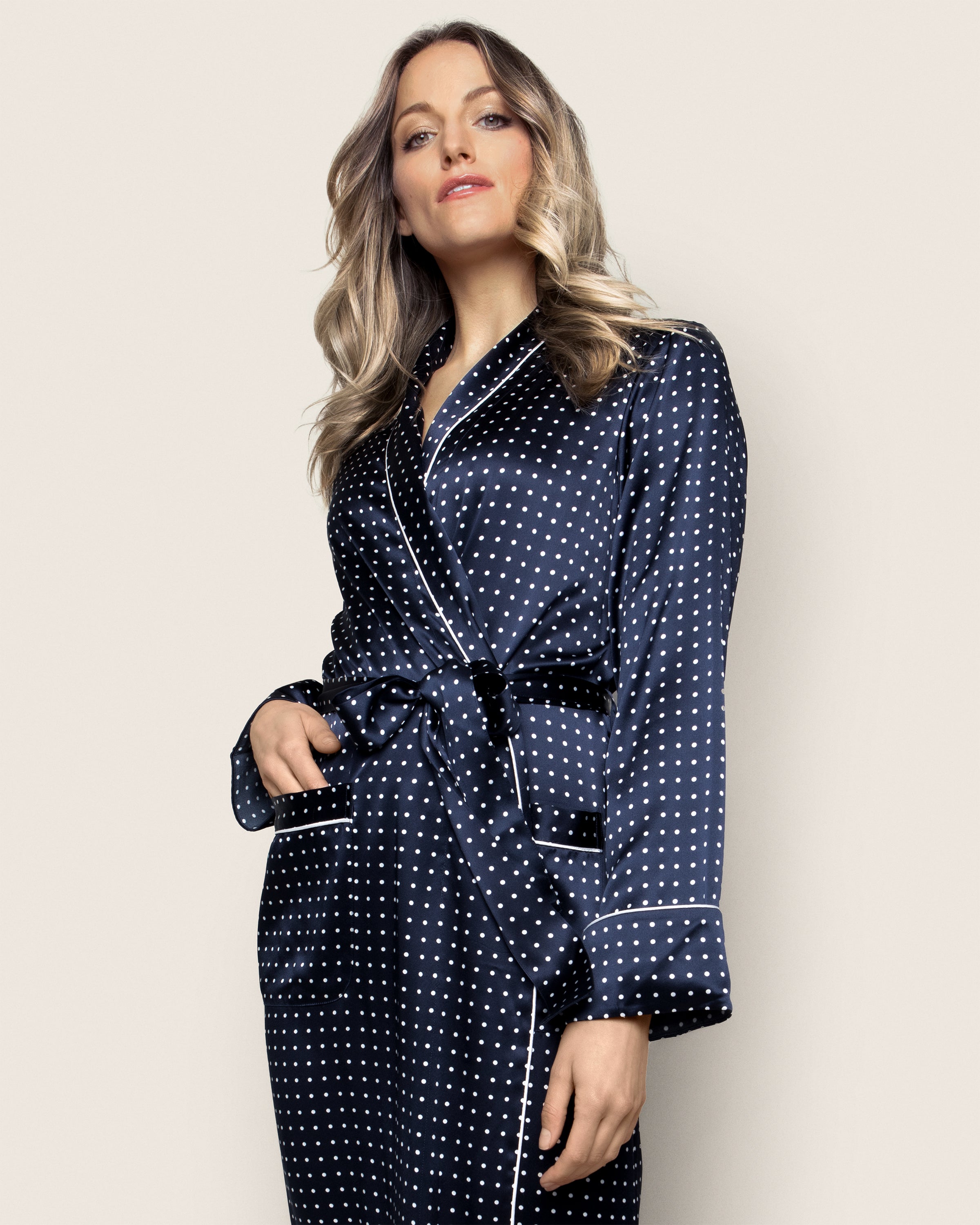 A person with long, wavy hair stands confidently in a Petite Plume Womens Silk Long Robe in Navy Polka Dot, tied at the waist, with a hand in one pocket. They pose elegantly against a beige background.