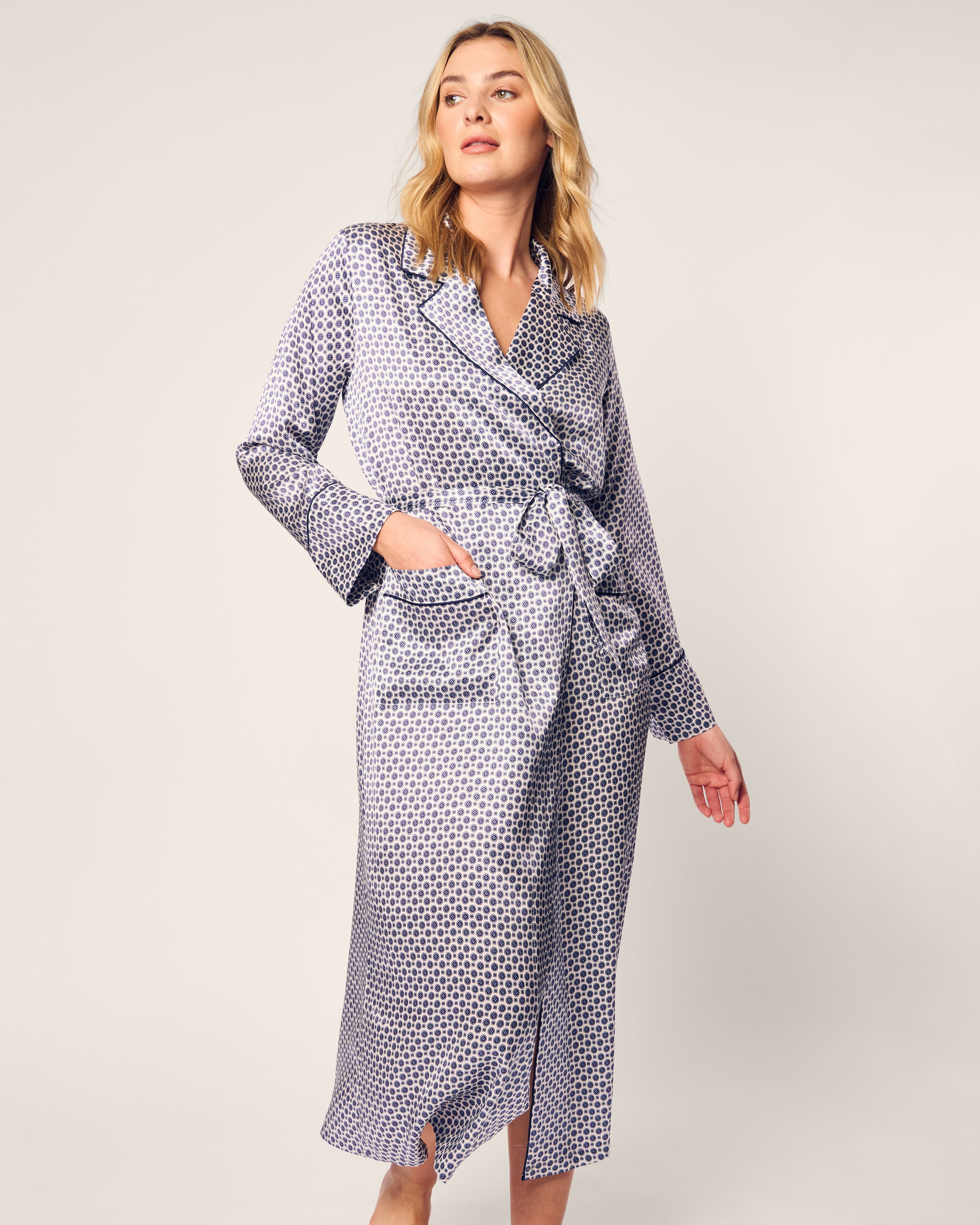 A person wears Petite Plumes Womens Silk Robe in Navy Foulard, featuring a geometric design in white and blue. Made from Mulberry silk, it has long sleeves and a waist tie, contrasting the plain light-colored background.