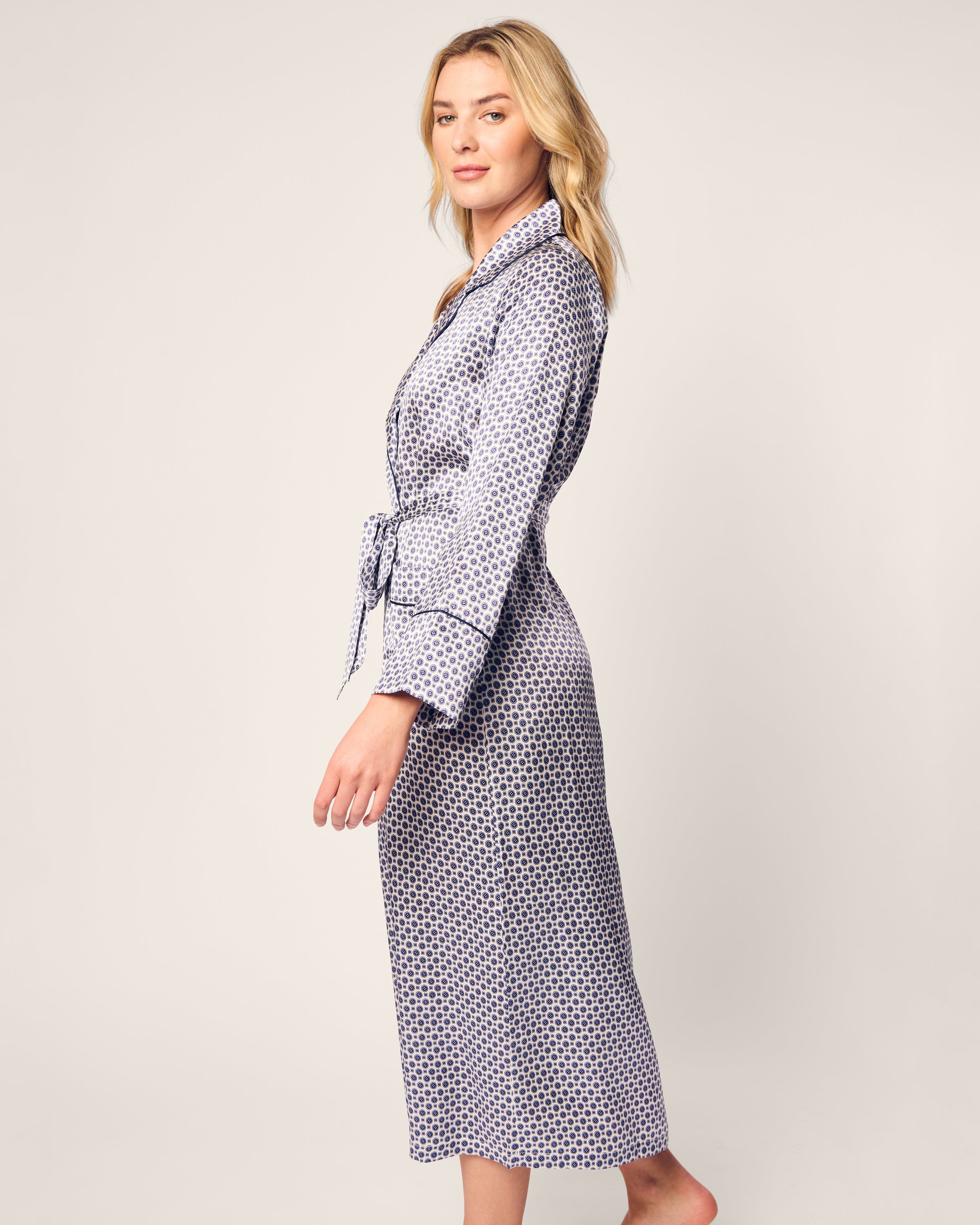 A person with long blond hair wears Petite Plumes Womens Silk Robe in Navy Foulard, resembling elegant sleepwear, complete with a belt. They stand barefoot on a light background, gazing to the side with a relaxed expression.