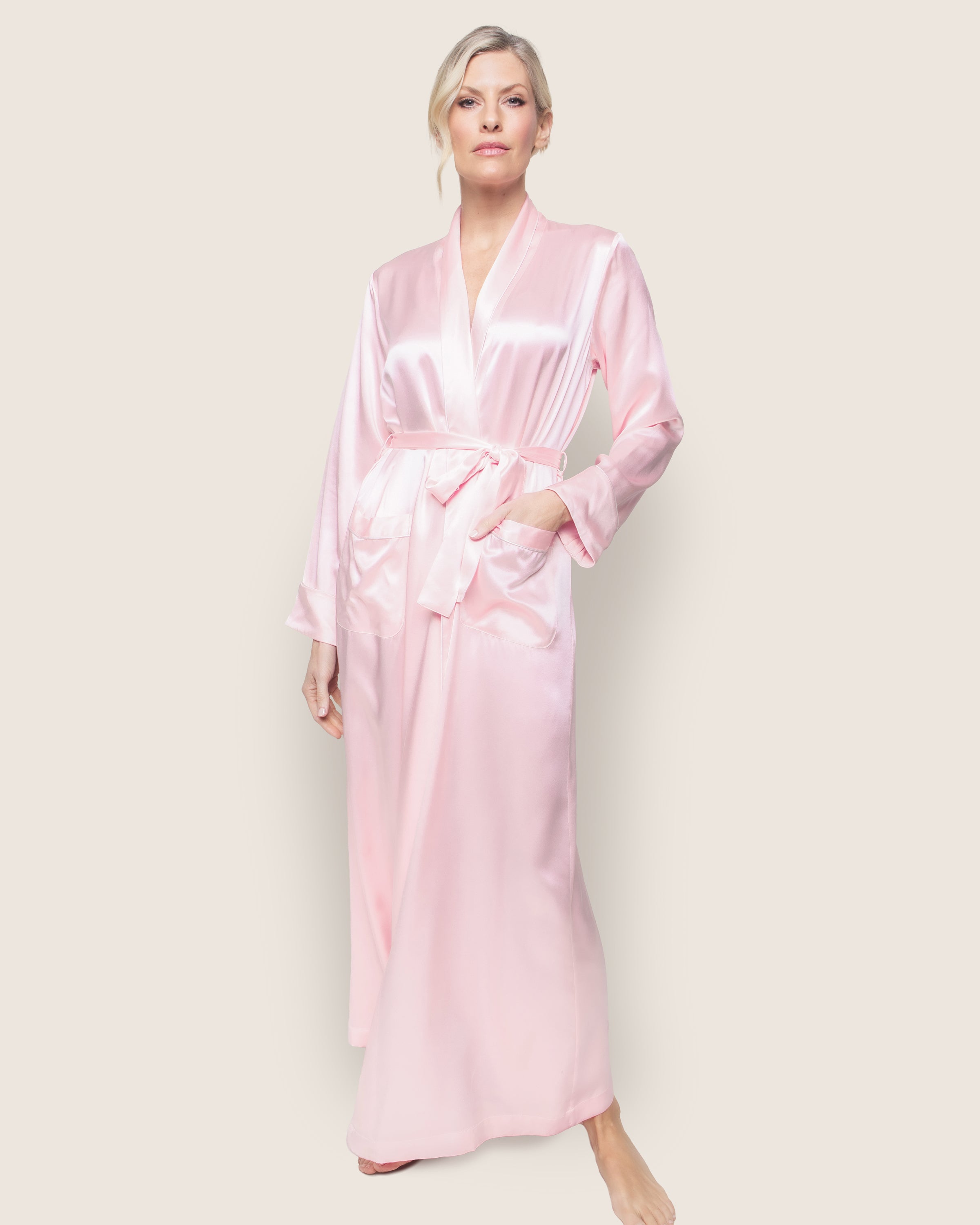 A person wears the Womens Silk Long Robe in Pink by Petite Plume, featuring a shiny texture and long sleeves. The elegantly crafted light pink robe with a tied belt epitomizes luxury sleepwear. They stand barefoot against a plain background, hair styled in an updo.