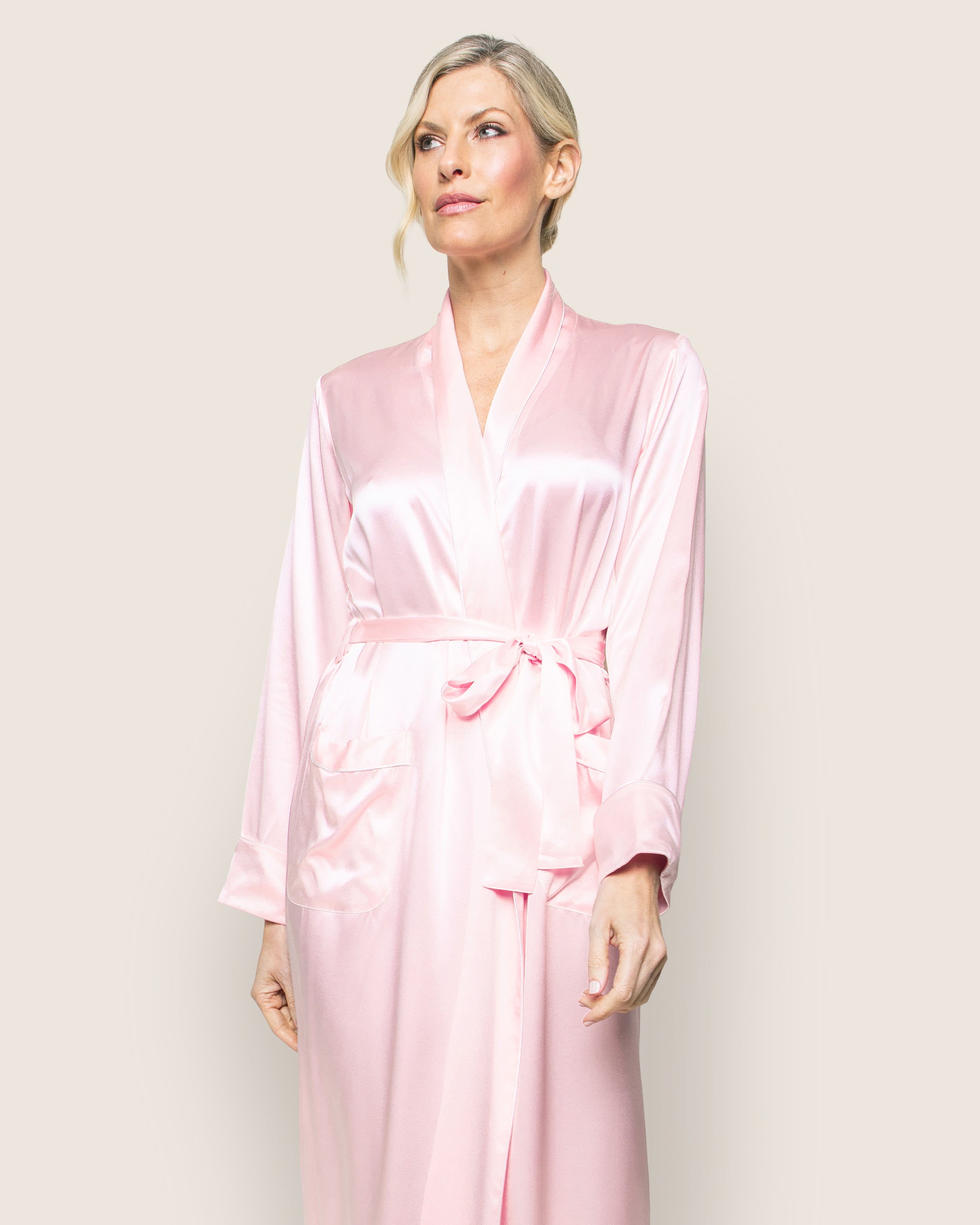 Against a plain backdrop, a person with short blonde hair embodies luxury sleepwear in Petite Plumes Womens Silk Long Robe in Pink. The silky fabric drapes elegantly, featuring a tied belt and front pockets.