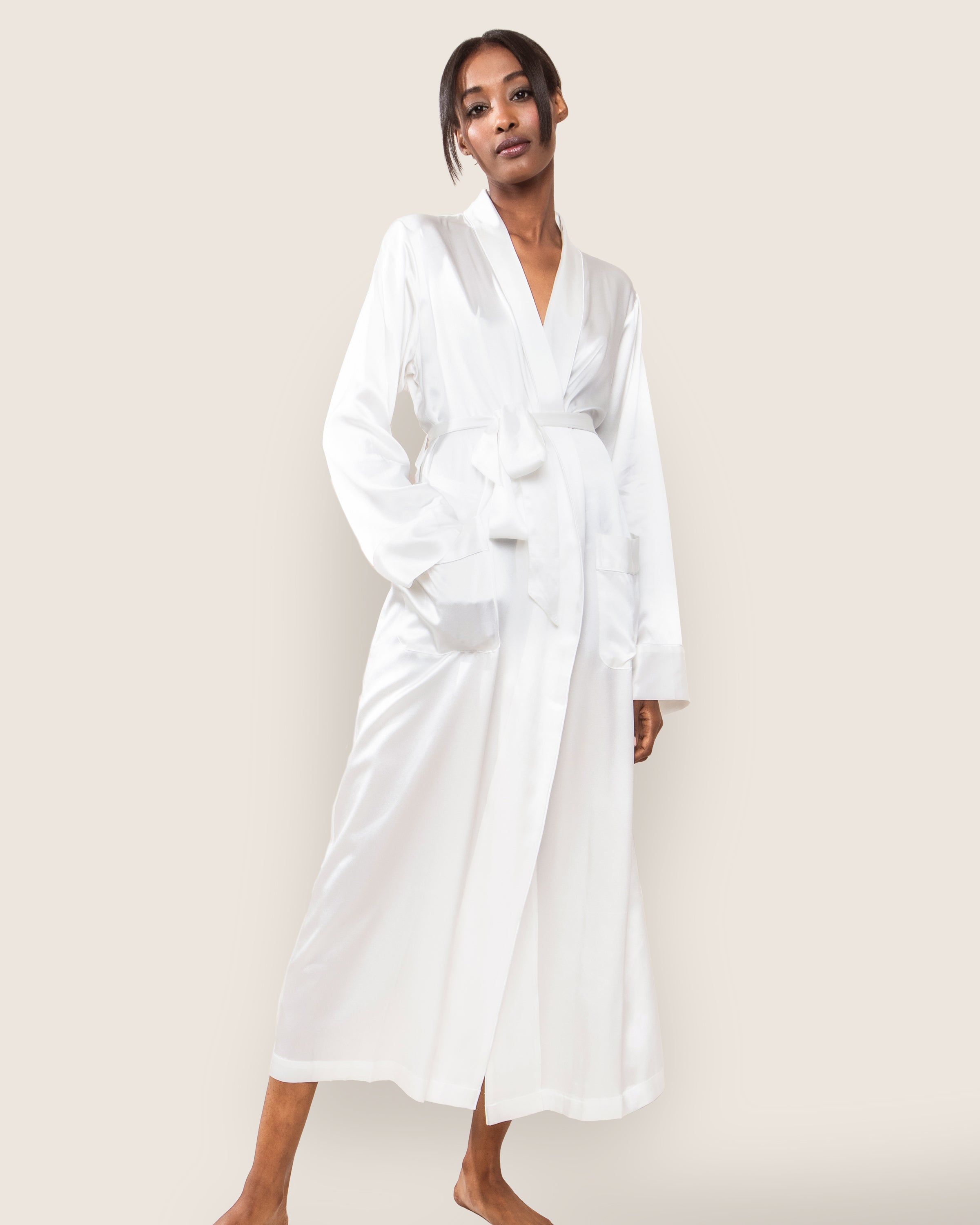 A person wearing the Women’s Silk Long Robe in White by Petite Plume, made of luxurious 22mm silk with long sleeves, a tie waist, and two front pockets, stands barefoot against a light background with short hair and looks directly at the camera, exuding timeless elegance.