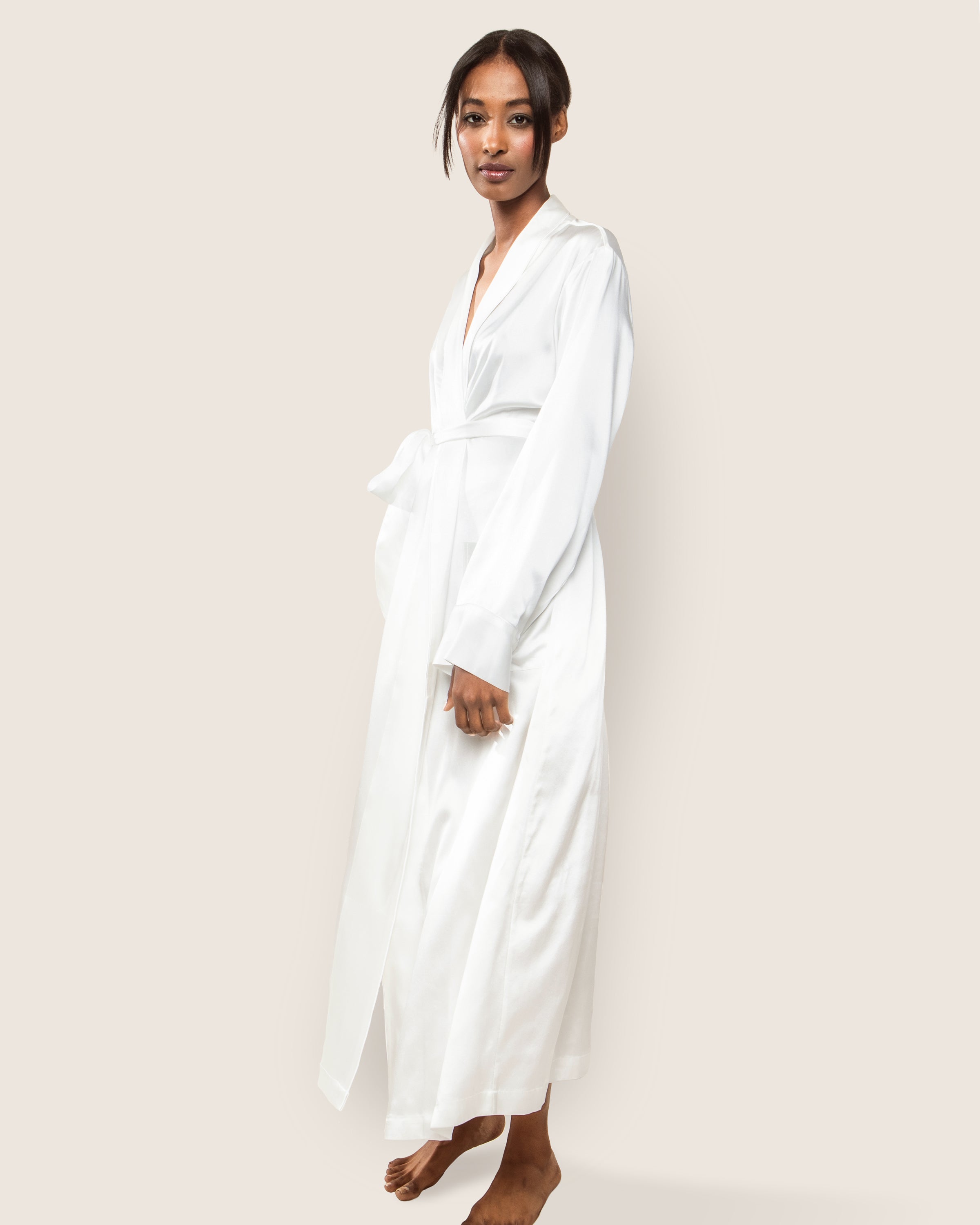 A person stands barefoot on a light background, wearing the Womens Silk Long Robe in White by Petite Plume, and looks at the camera with a calm expression.