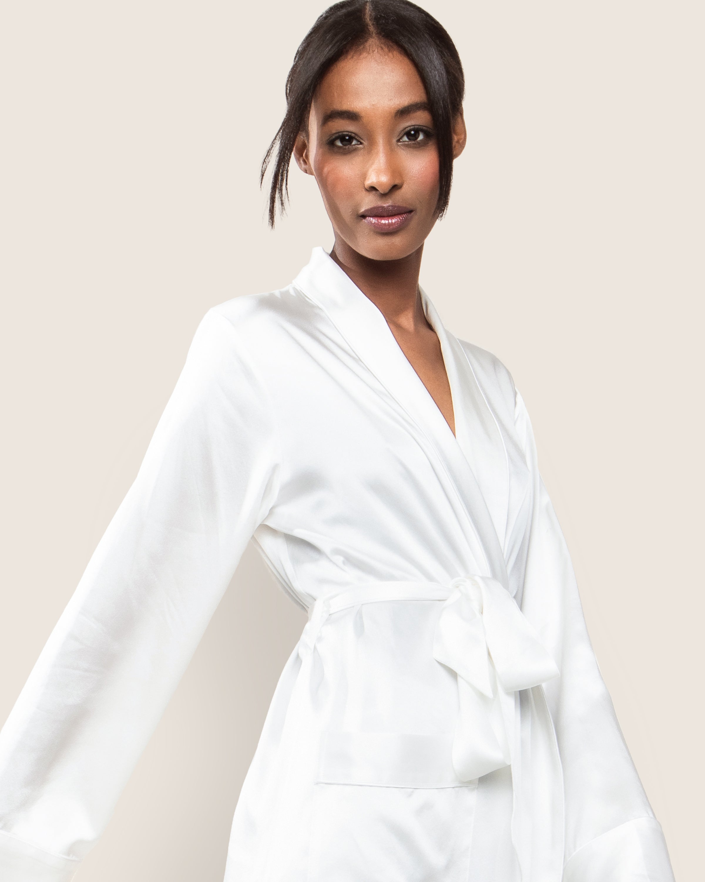 A woman poses elegantly in a Petite Plume Womens Silk Long Robe in White, featuring long sleeves and a tied waist, against a plain beige background.