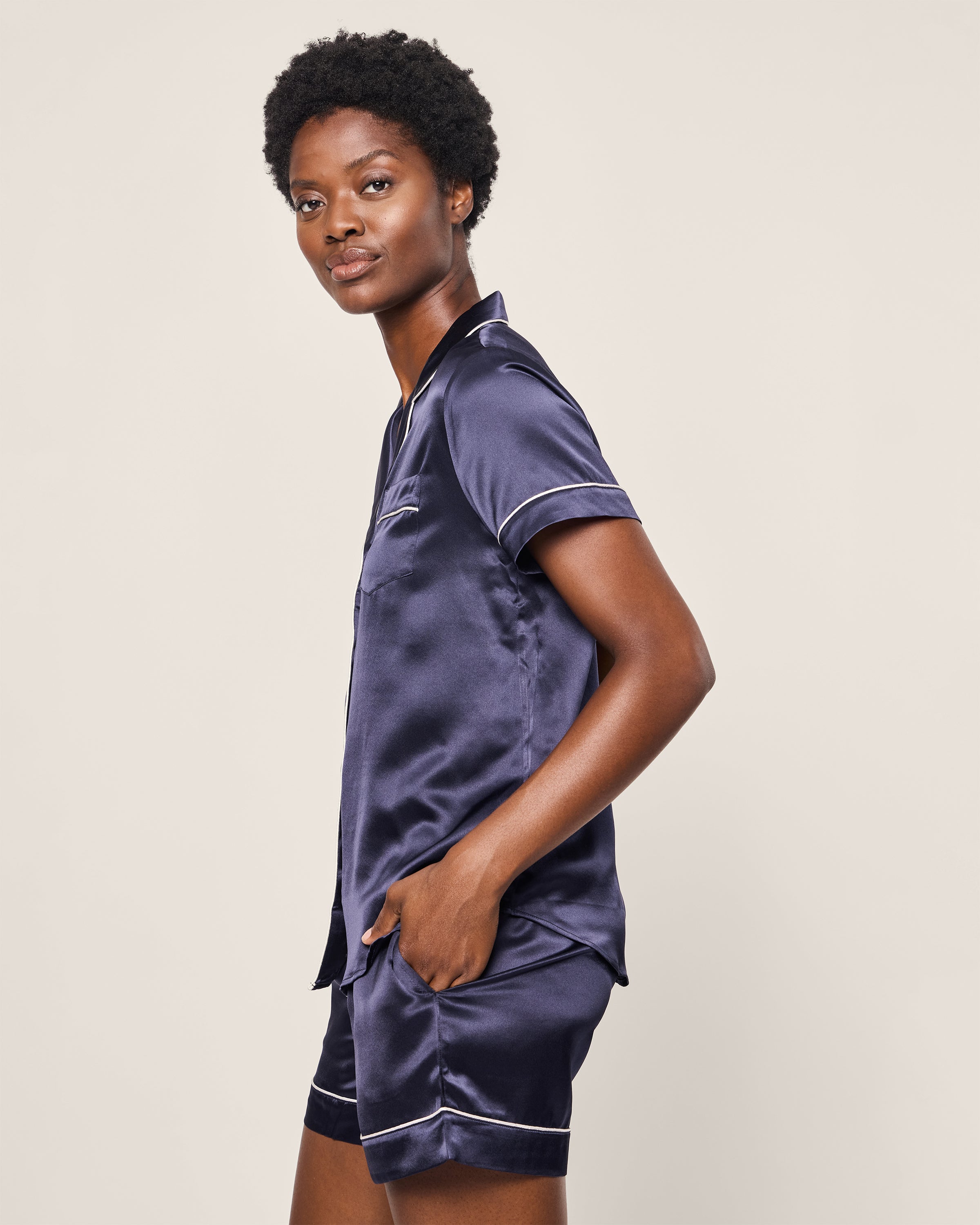 Against a plain backdrop, a person with short curly hair wears the Womens Silk Short Set in Navy by Petite Plume, featuring white piping. Their hand casually rests in the pajama shorts pocket.