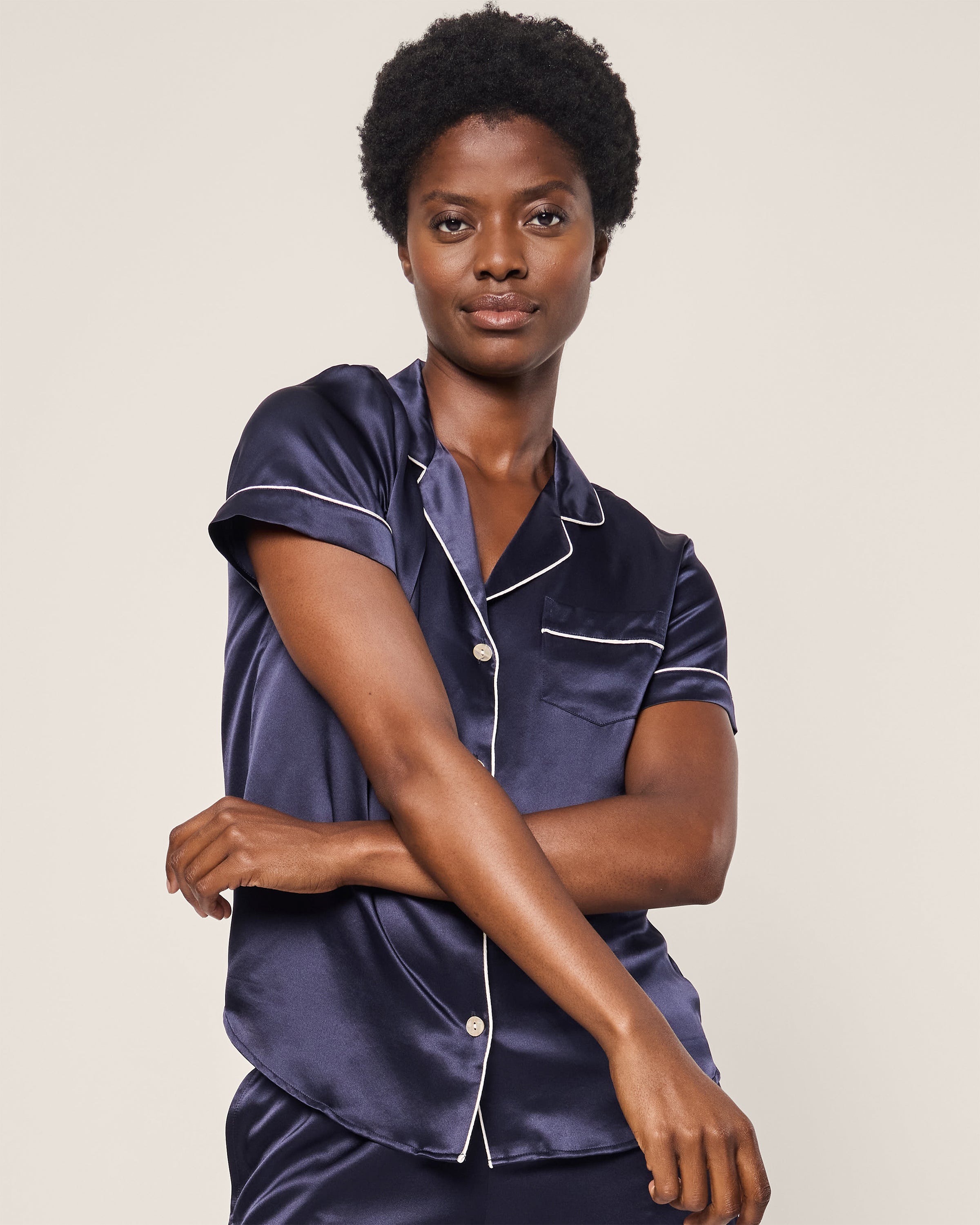 A person with short curly hair, wearing Petite Plumes Womens Silk Short Set in Navy, exudes luxurious elegance. Against a neutral background, their gently crossed arms embody the exquisite allure of silk sleepwear.