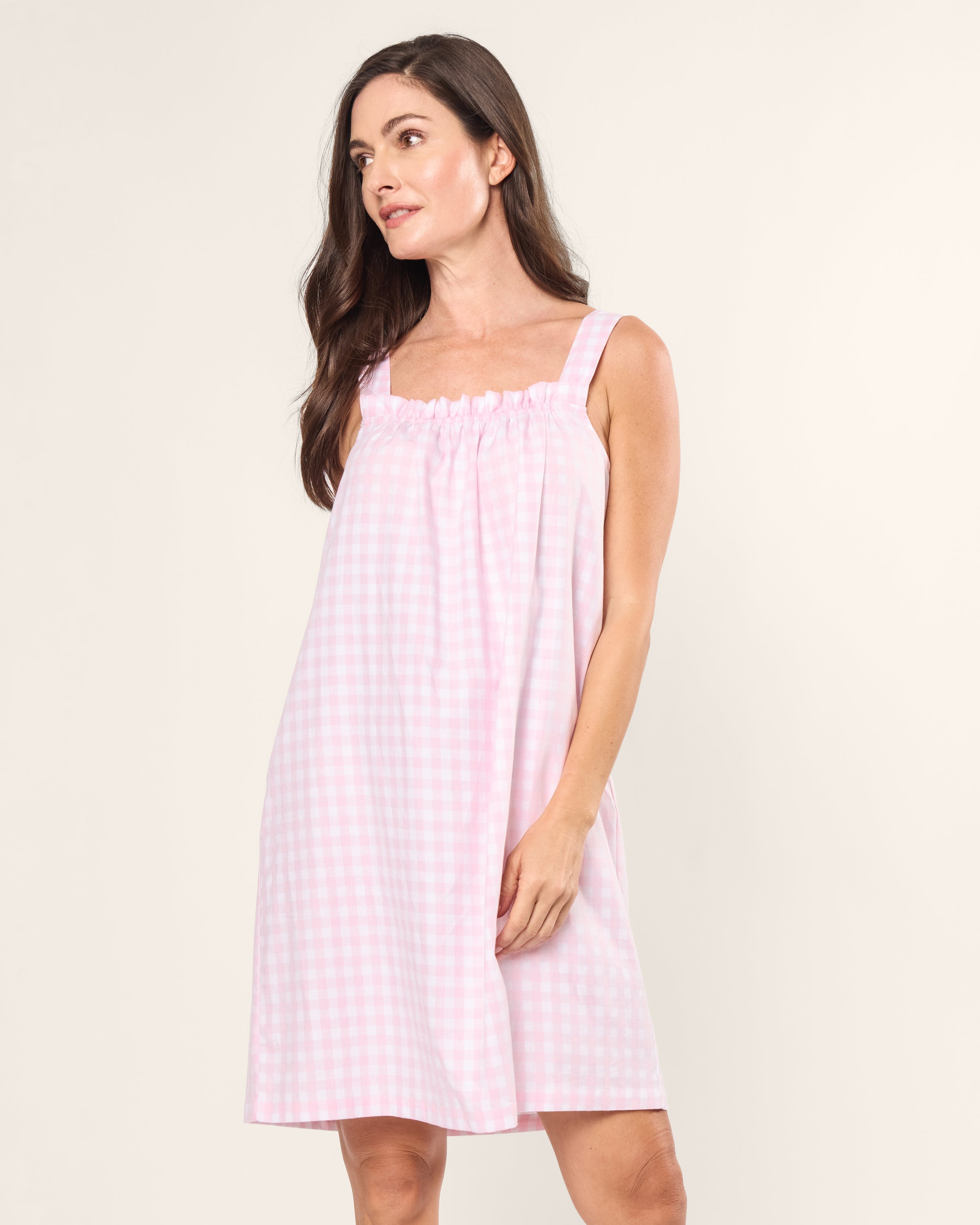 A woman with long dark hair models a sleeveless pink gingham dress against a plain light background, slightly smiling to her left. Her right hand gently touches the dresss side, echoing the elegance of the Petite Plume Womens Twill Annabelle Nightgown in Pink Gingham.