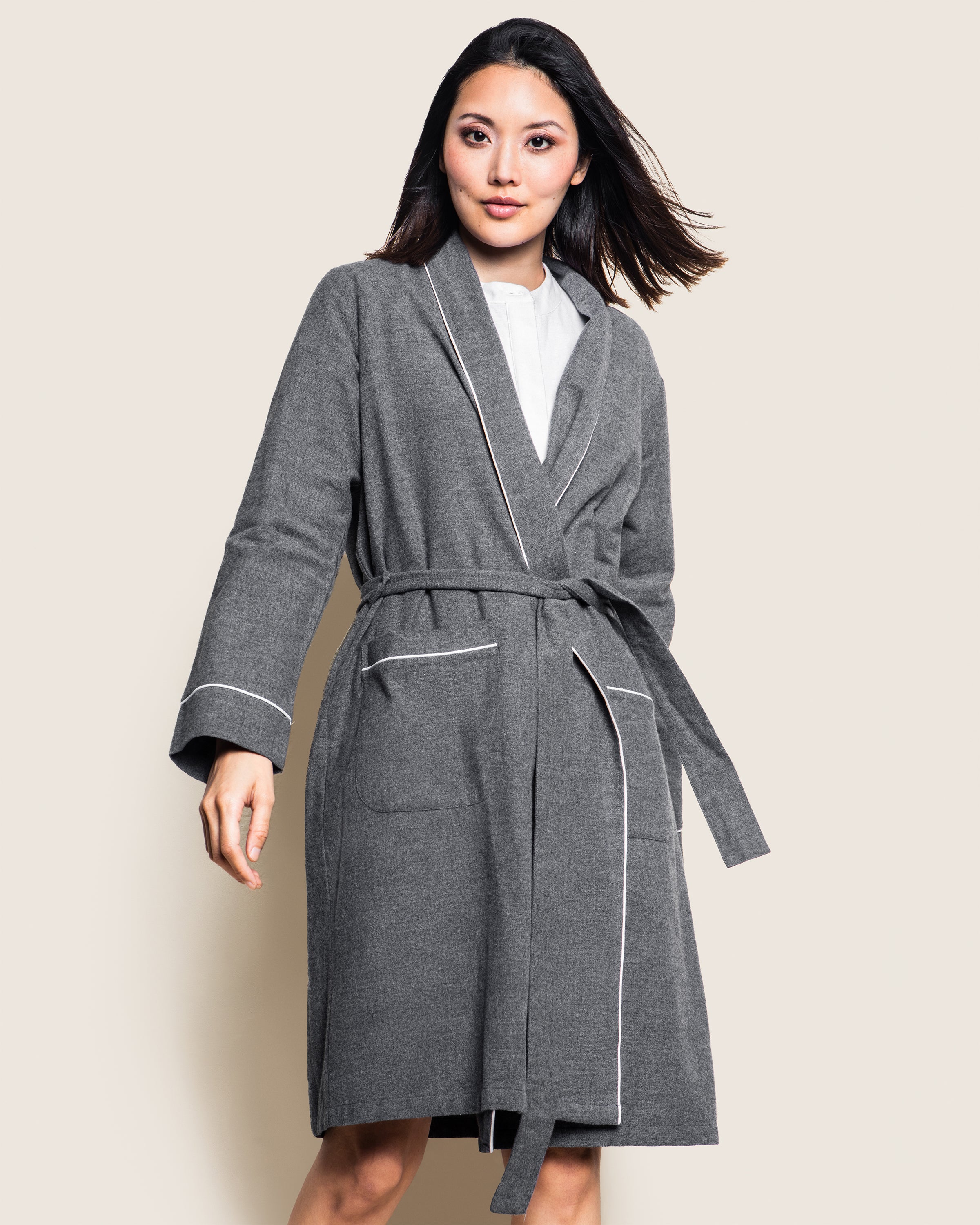 A person wearing a Womens Flannel Robe in Grey by Petite Plume, made of 100% cotton with white trim, stands against a plain background. The luxurious robe includes a belt and large pockets, while the individual faces forward with a slight smile.