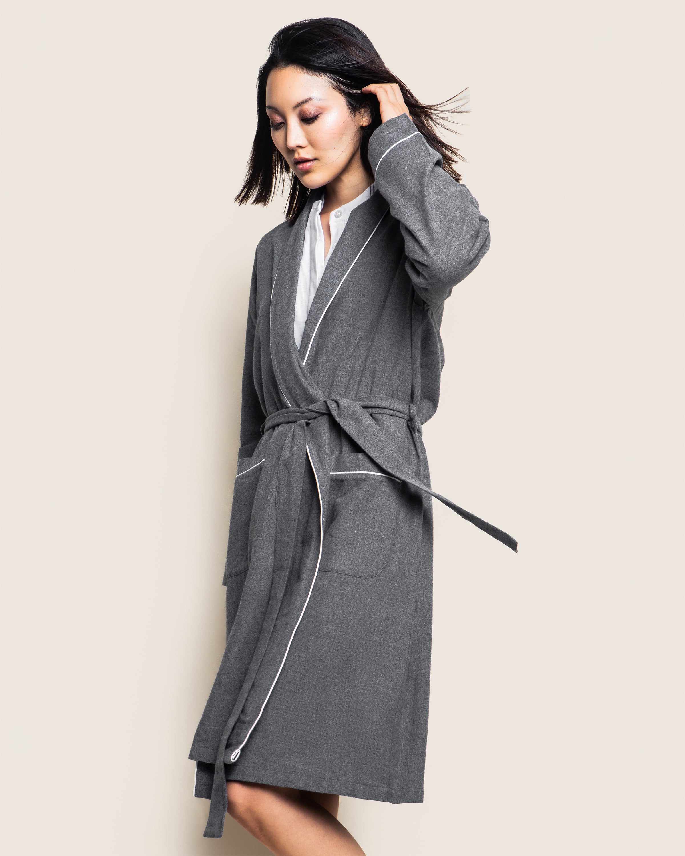 A person wearing an elegant Womens Flannel Robe in Grey by Petite Plume stands against a light background. One hand touches their hair while the other rests by their side, with the luxurious robe tied at the waist, revealing a white shirt underneath.