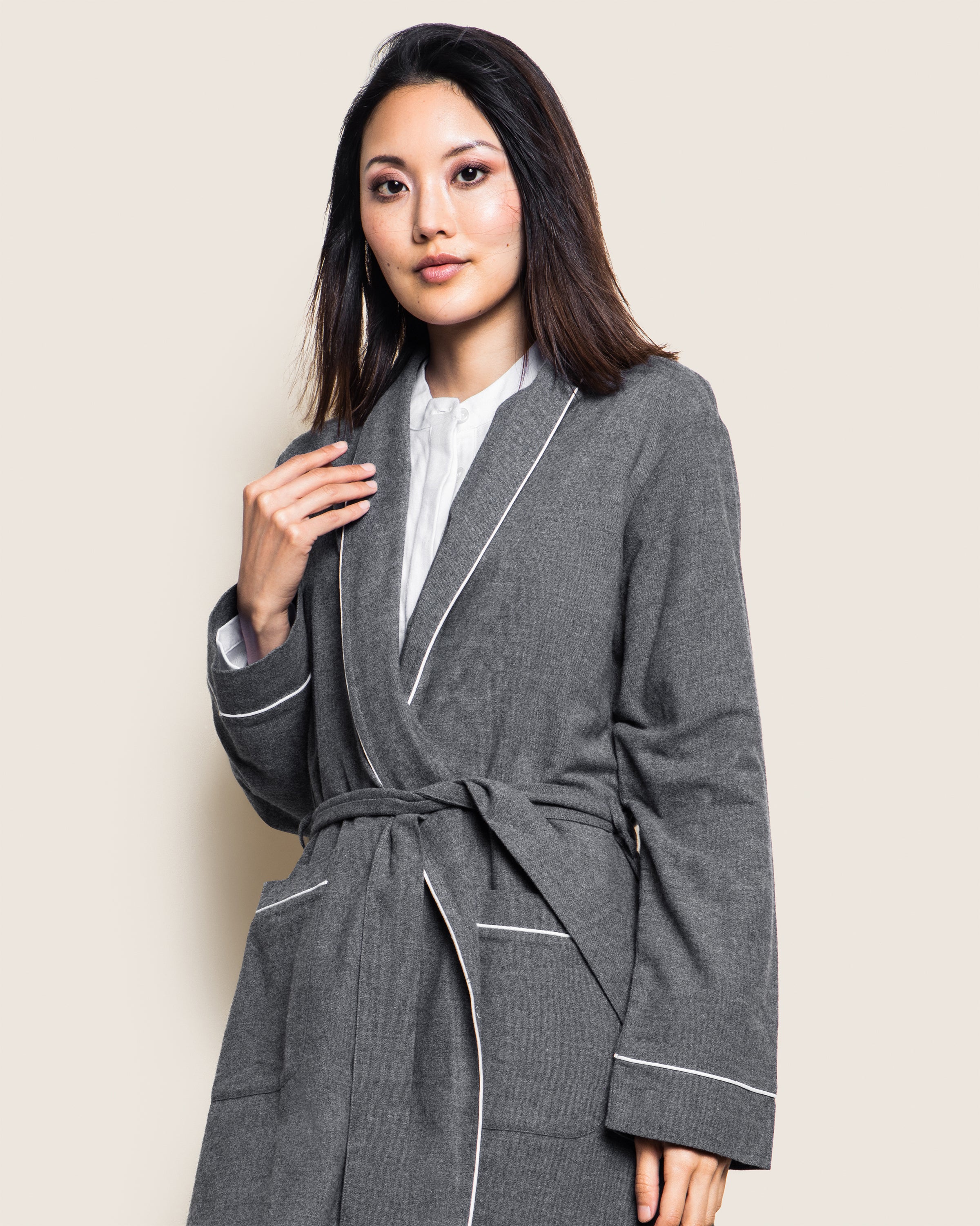 A person models the Petite Plume Womens Flannel Robe in Grey, featuring white piping, against a light beige backdrop. With medium-length dark hair and a white shirt underneath, they look directly at the camera with confidence.