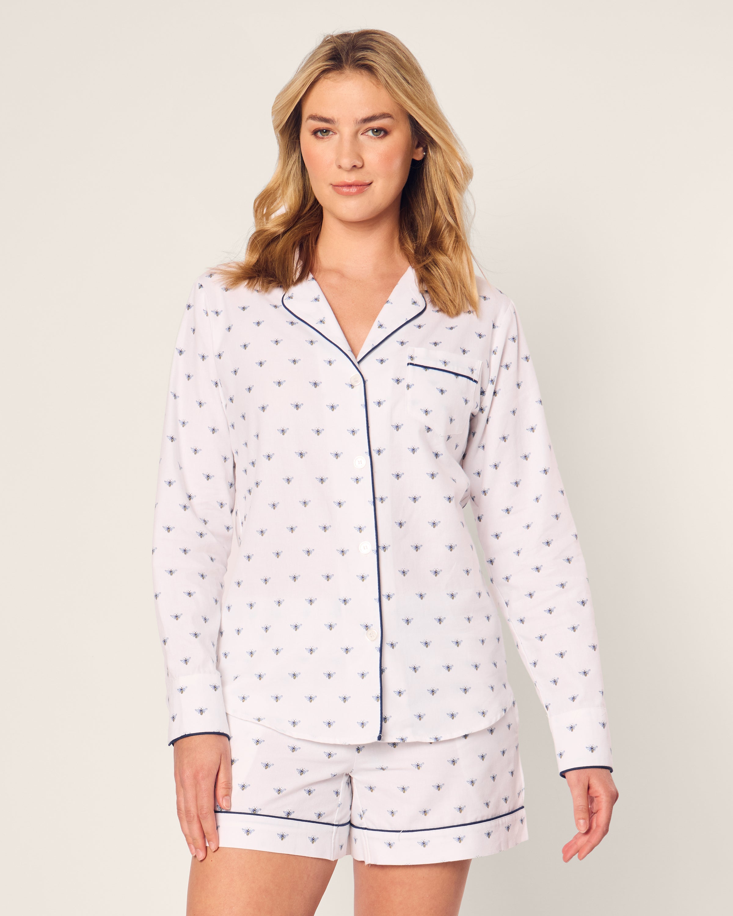 The woman models Petite Plumes Womens Twill Long Sleeve Short Set in Buzzing Bees, featuring a white pajama with small blue bee patterns, long sleeves, and a dark blue piped collar. She pairs it with matching shorts against a plain white backdrop.