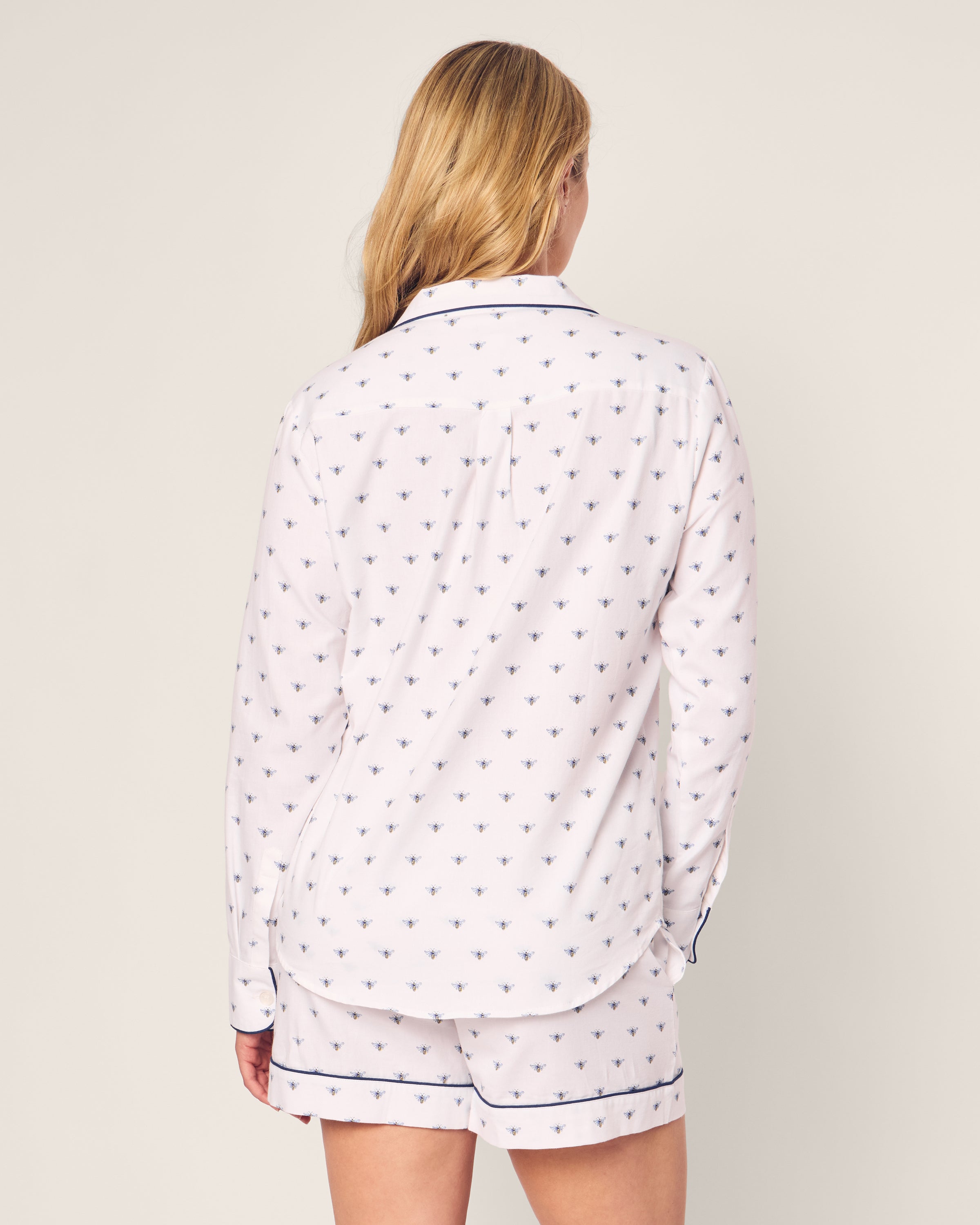 A person with long blonde hair is shown from the back wearing Petite Plumes Womens Twill Long Sleeve Short Set in Buzzing Bees. The luxurious sleepwear features a white top with long sleeves and a collar, while the shorts have an elastic waistband. The background is a plain light color.