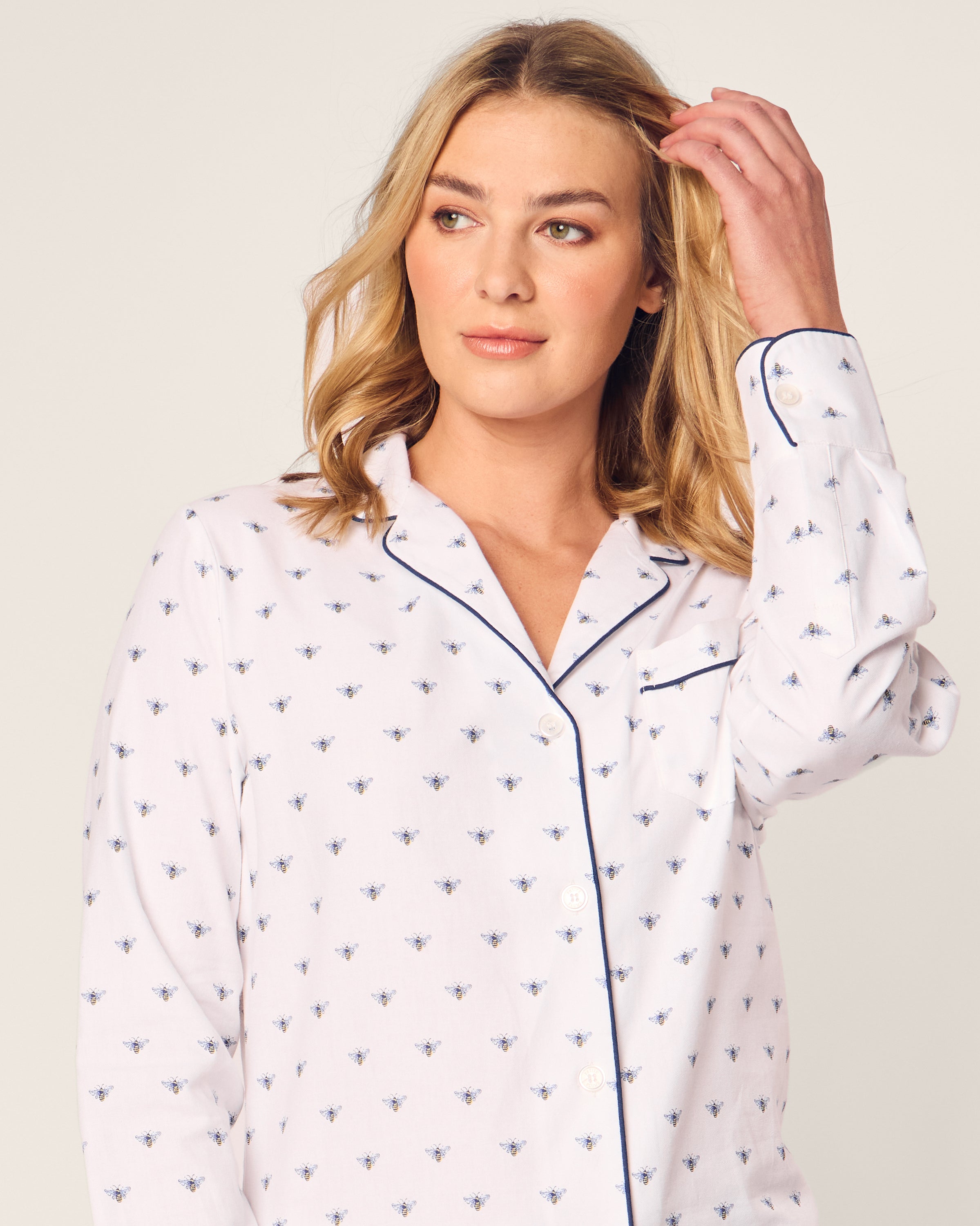 A person with shoulder-length blonde hair, wearing Petite Plumes Womens Twill Long Sleeve Short Set in Buzzing Bees, touches their hair while gazing to the side against a plain, light-colored background. The luxurious white sleepwear features small bees and dark piping.