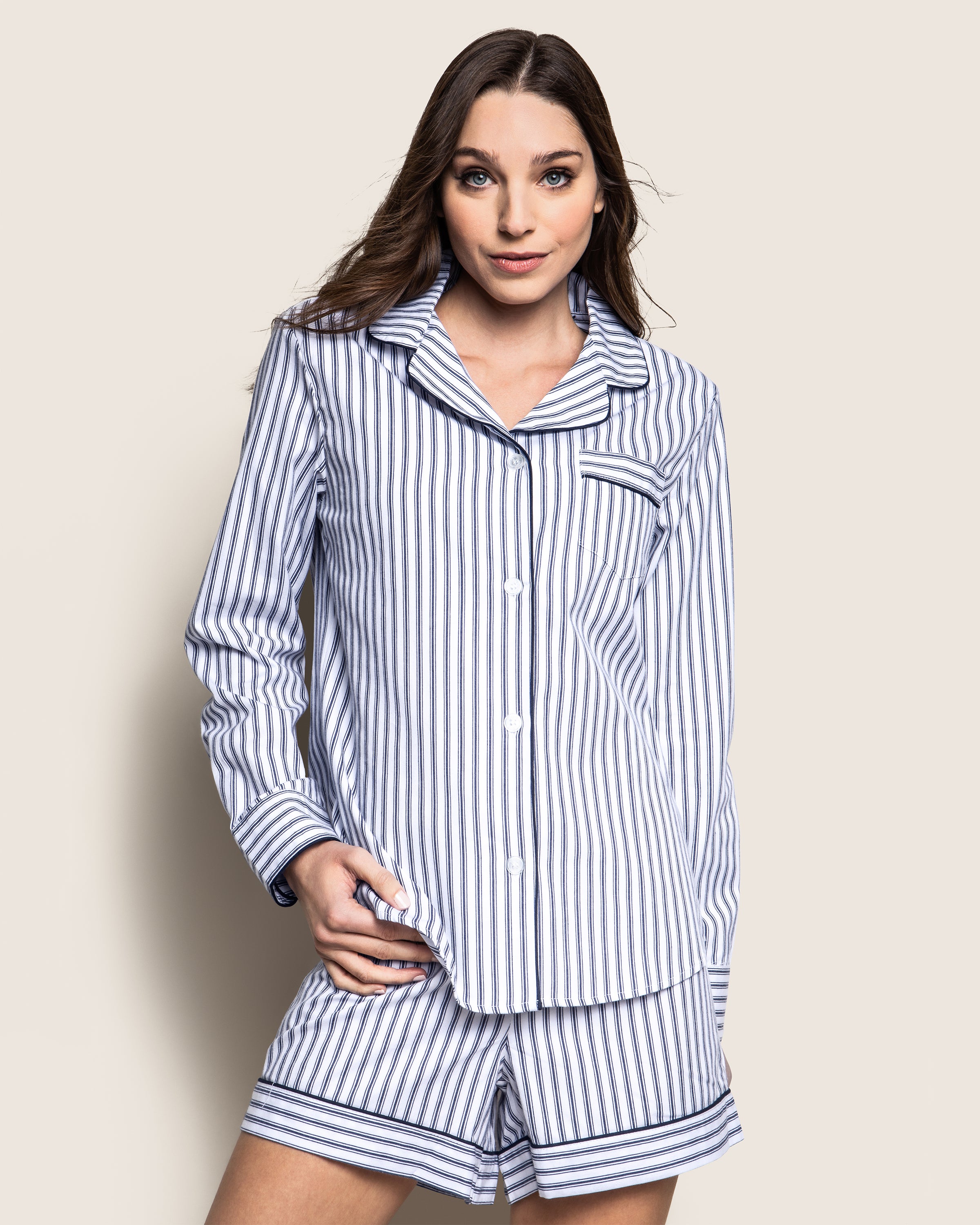 A person with long brown hair poses against a light background, wearing Petite Plumes Womens Twill Long Sleeve Short Set in Navy French Ticking—a 100% cotton outfit featuring elegant white and light blue vertical stripes on a button-up top and shorts.