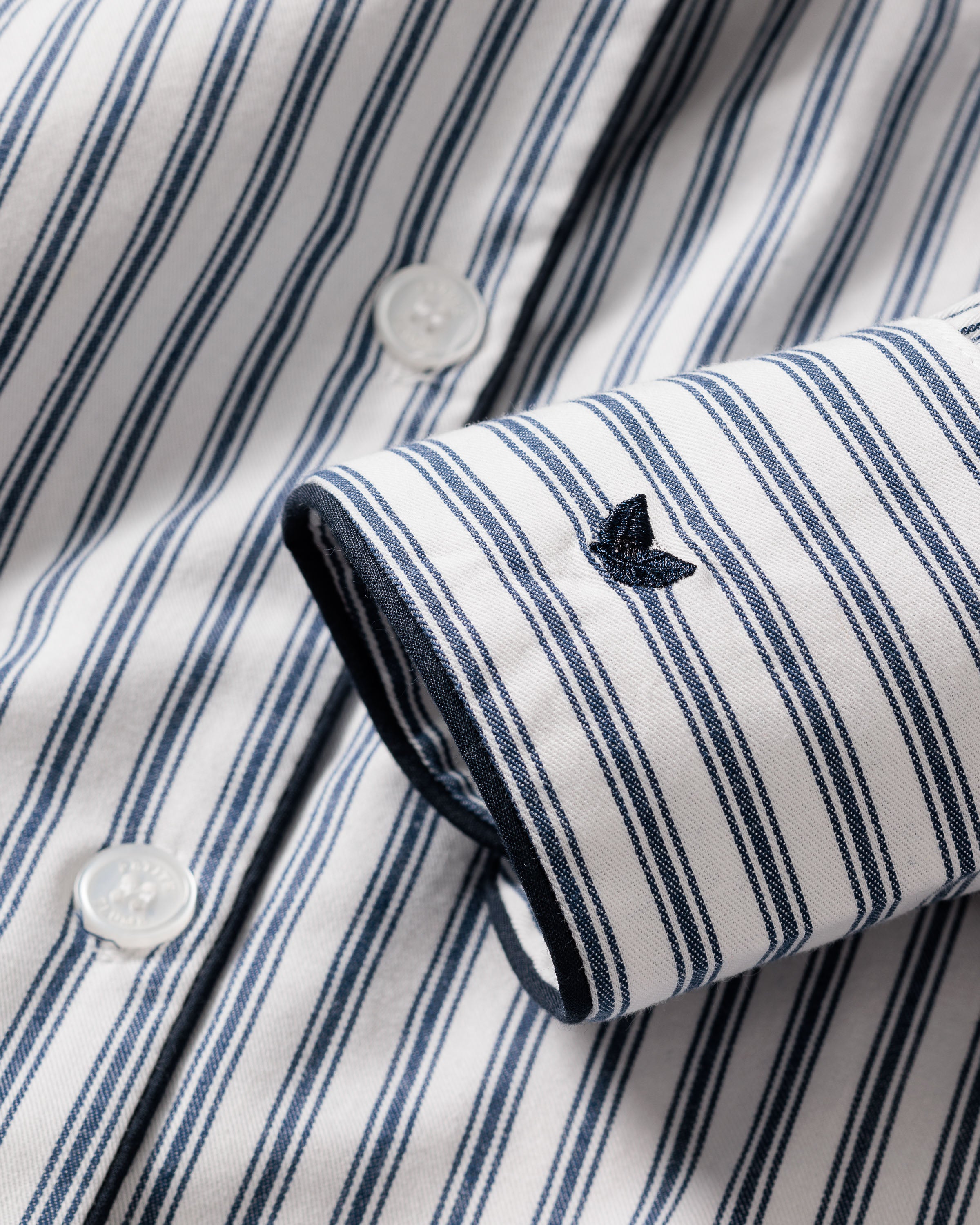 A close-up of Petite Plume’s Womens Twill Long Sleeve Short Set in Navy French Ticking shows a white shirt with navy stripes, white buttons, and a cuff embroidered in navy on smooth cotton fabric.