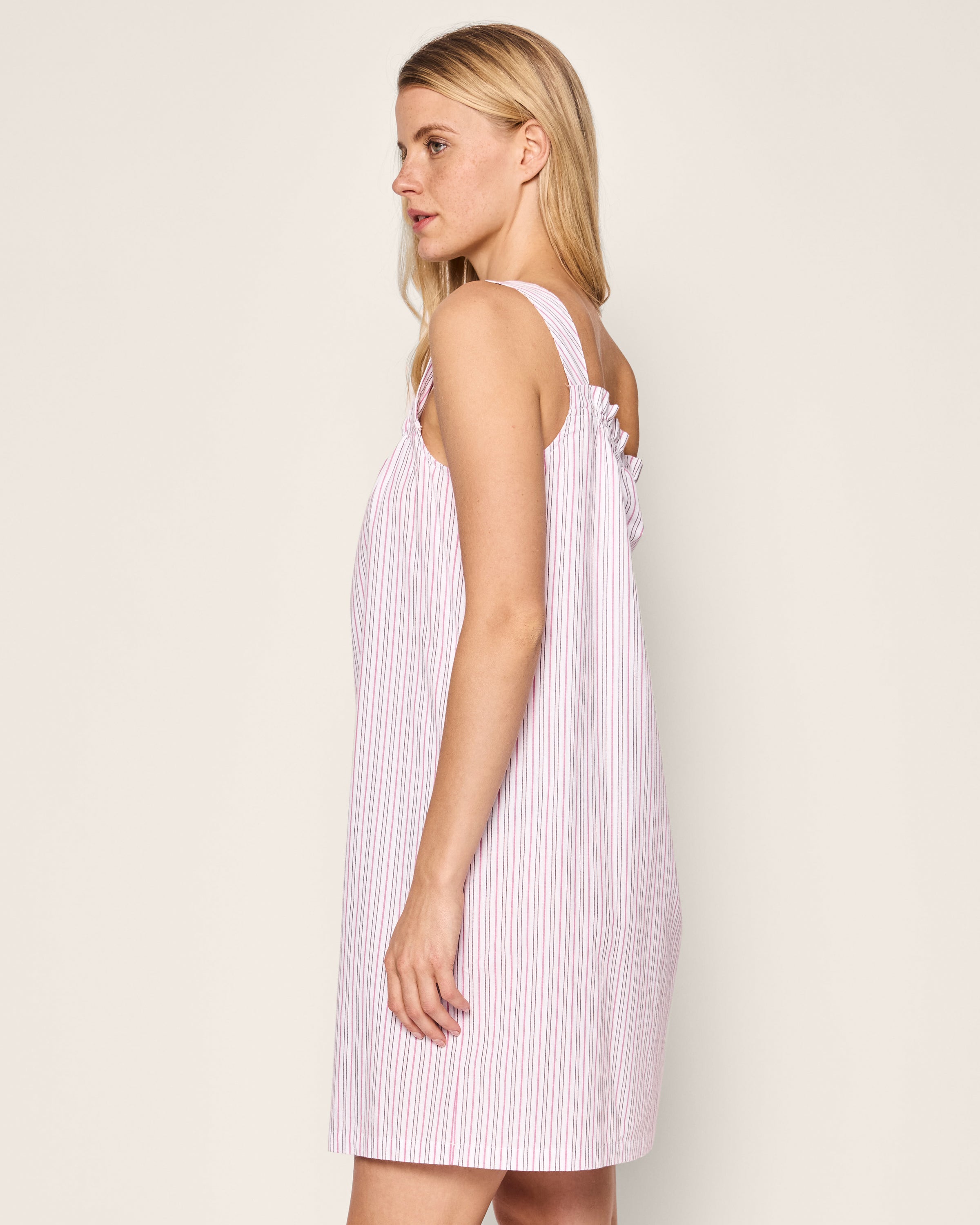 A person with long blonde hair wears a sleeveless, pink and white striped Petite Plume Womens Twill Annabelle Nightgown in Blush Boulevard. They stand sideways, slightly facing right. The nightgown boasts ruffled straps and a loose fit, set against a light background.