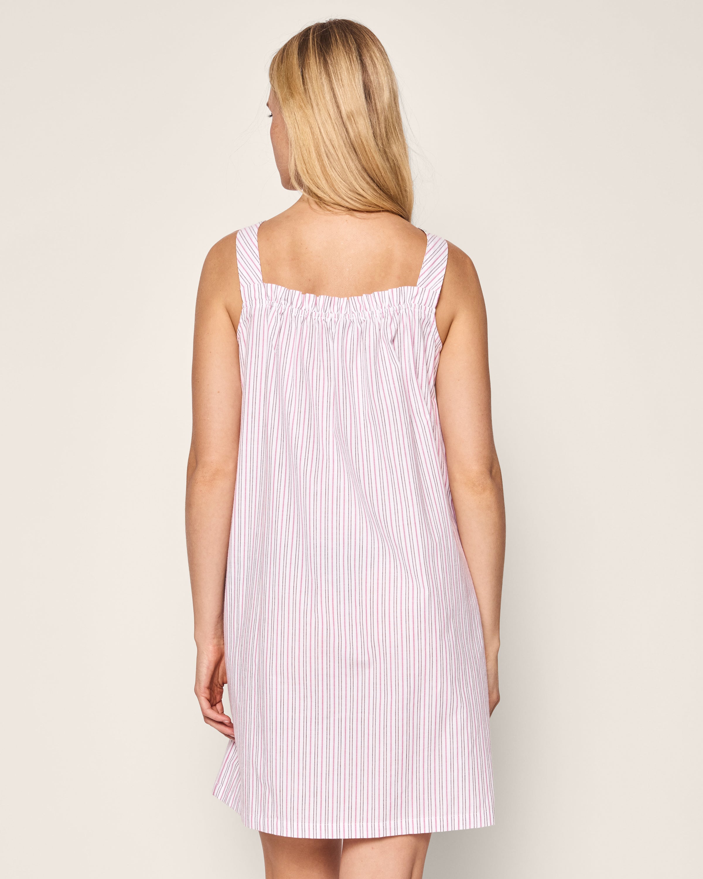 A blonde woman wearing a sleeveless knee-length Petite Plume Womens Twill Annabelle Nightgown in Blush Boulevard stands with her back to the camera against a plain beige background, showcasing the elegance of luxury sleepwear.