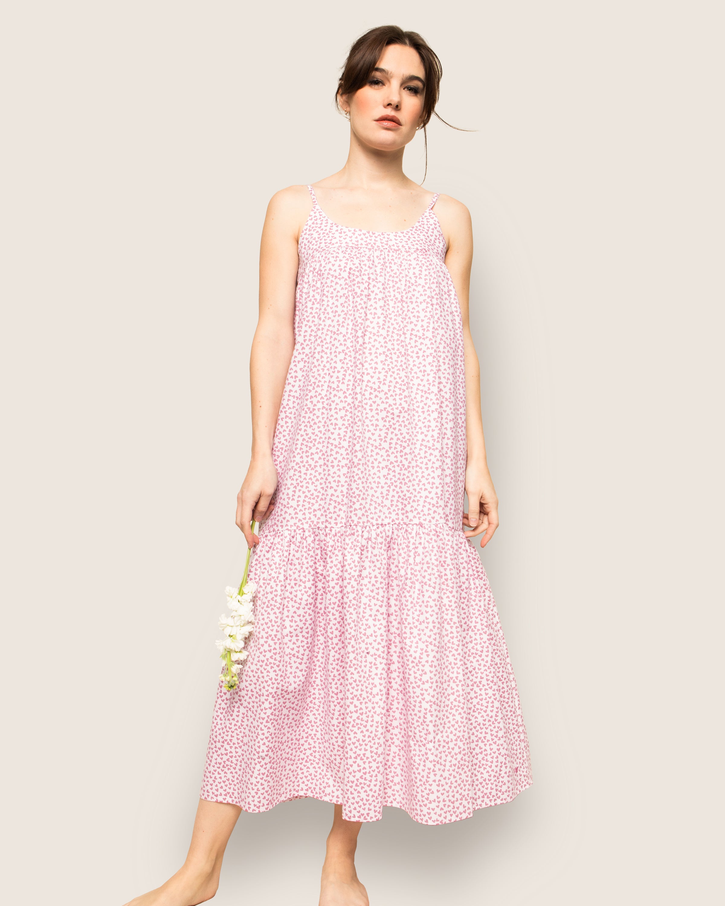 A woman in Petite Plumes Womens Twill Chloé Nightgown in Sweethearts, resembling luxury sleepwear, holds white flowers. She stands barefoot against a light beige background, with her hair tied back and loose strands framing her face, conveying subtle elegance and comfort.