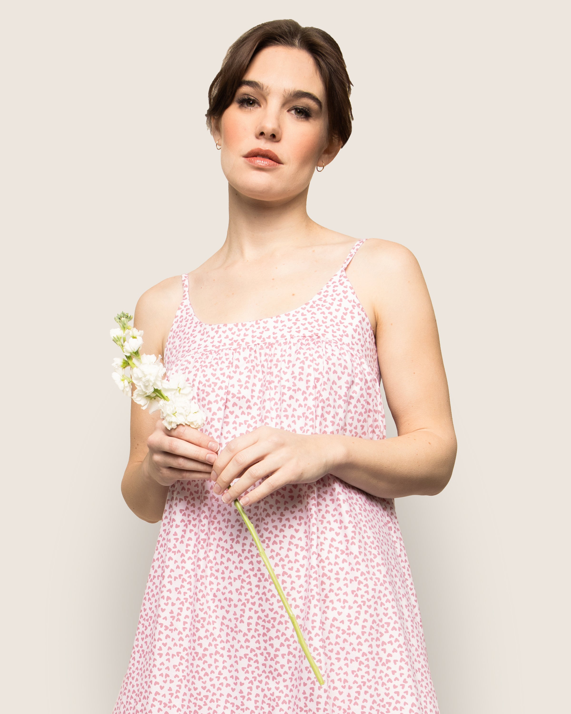 A woman in a Petite Plume Womens Twill Chloé Nightgown in Sweethearts, holding a single white flower, stands against a plain beige background, gazing slightly upward.