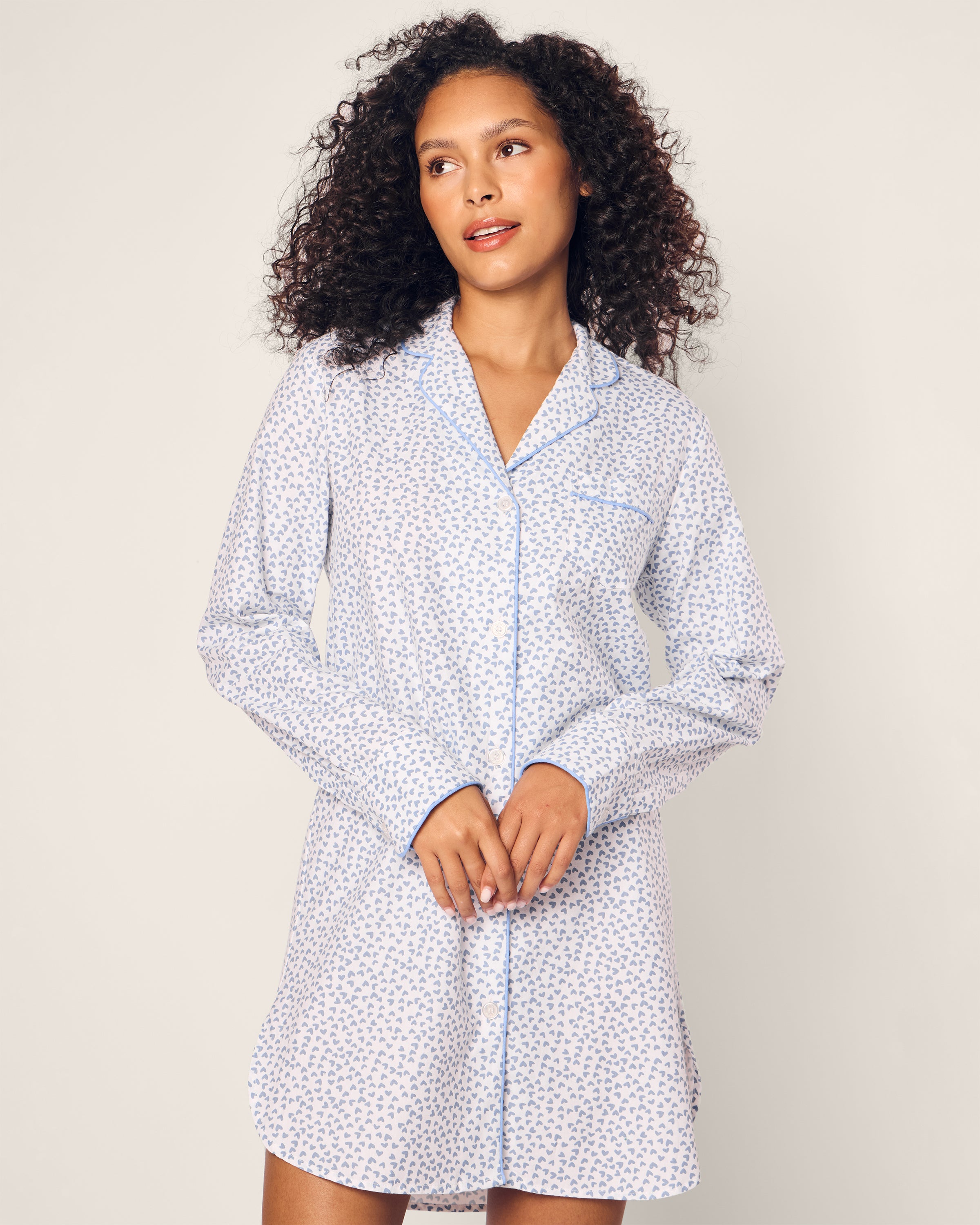 A person with curly hair wears the Womens Twill Nightshirt in Bluehearts by Petite Plume, featuring a small floral pattern. They look to the side with hands gently clasped, set against a minimalist light background.