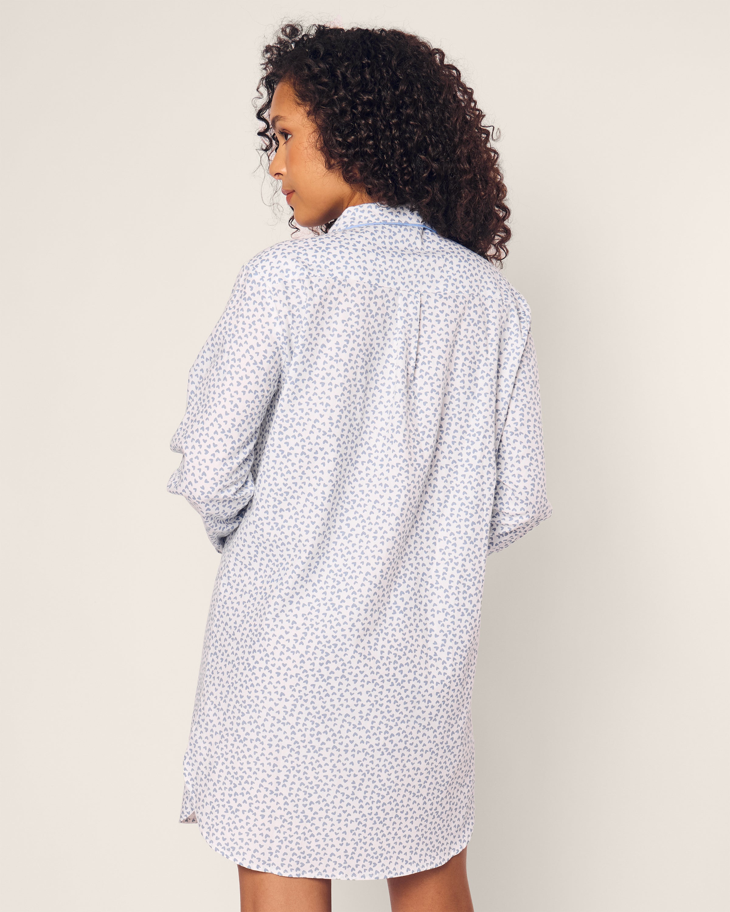 Featured is a woman with curly hair wearing a Petite Plume Womens Twill Nightshirt in Bluehearts, facing away from the camera against a minimalist background.