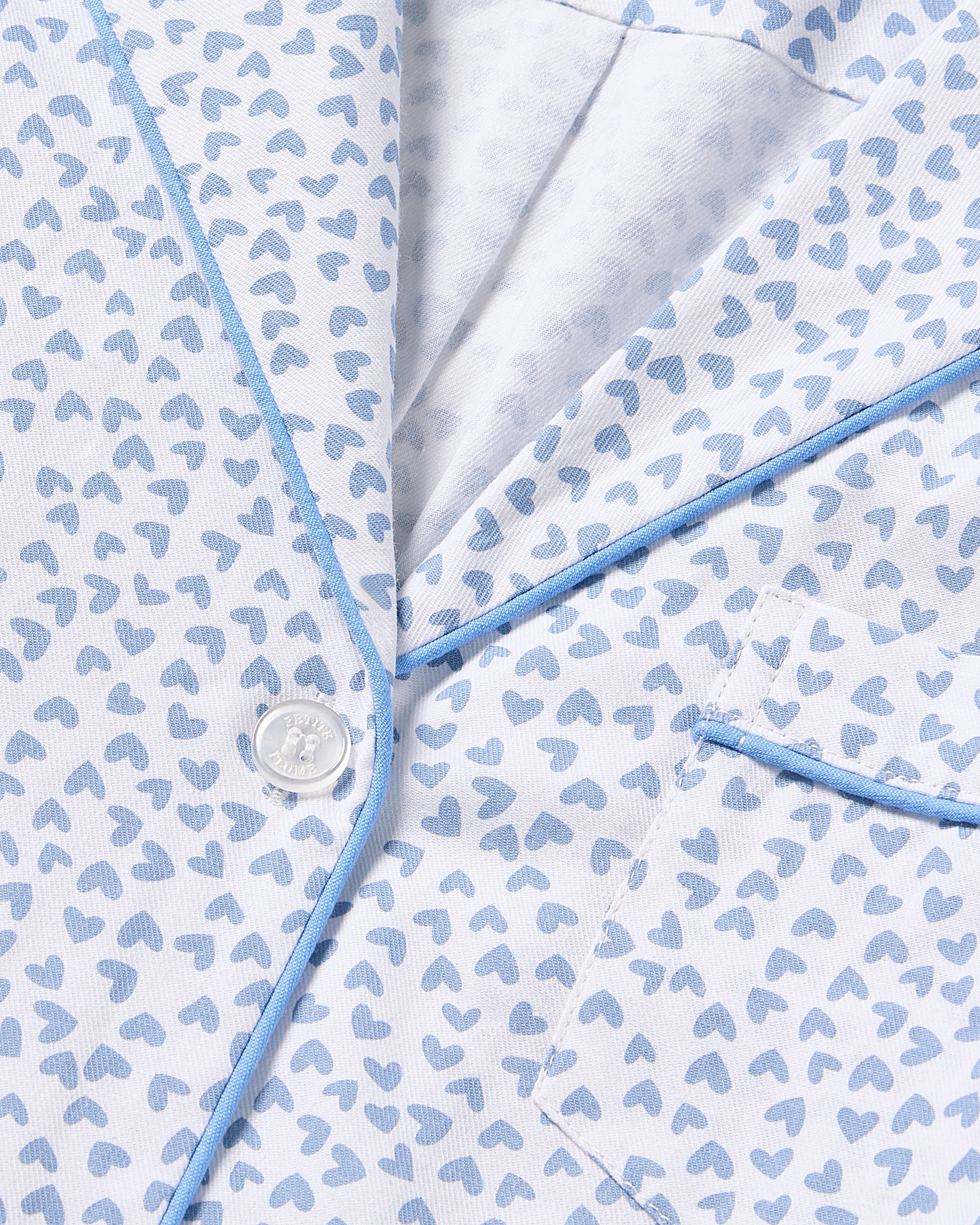 A close-up of Petite Plumes Womens Twill Nightshirt in Bluehearts reveals a minimalist white fabric with light blue hearts, featuring blue piping, a button, and a pocket. The lightweight cotton twill garment appears soft.