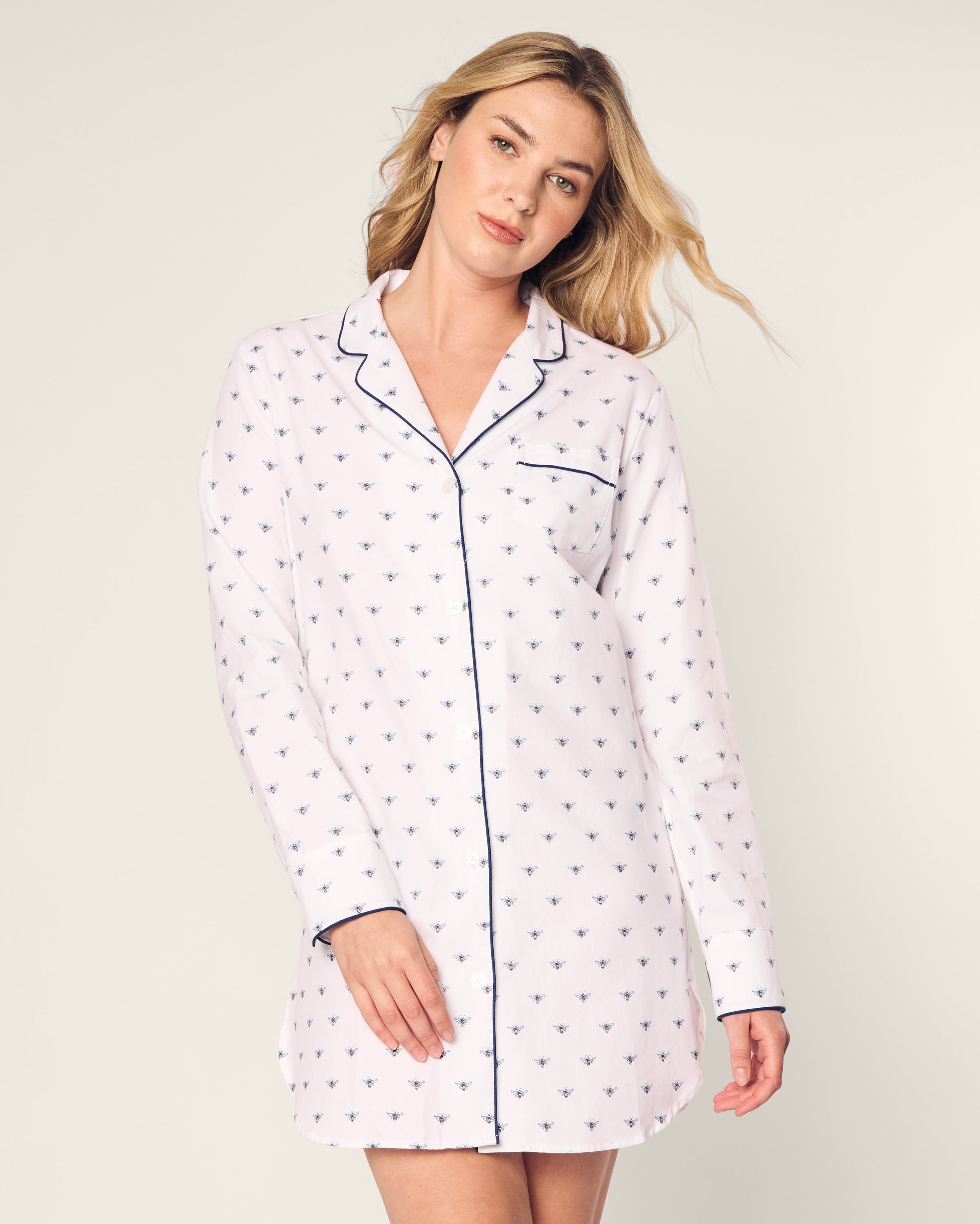 A person with long, wavy hair is wearing the Petite Plume Womens Twill Nightshirt in Buzzing Bees. The nightshirt is white with small dark bee patterns, featuring a collar and dark piping accents. They are standing against a plain background.