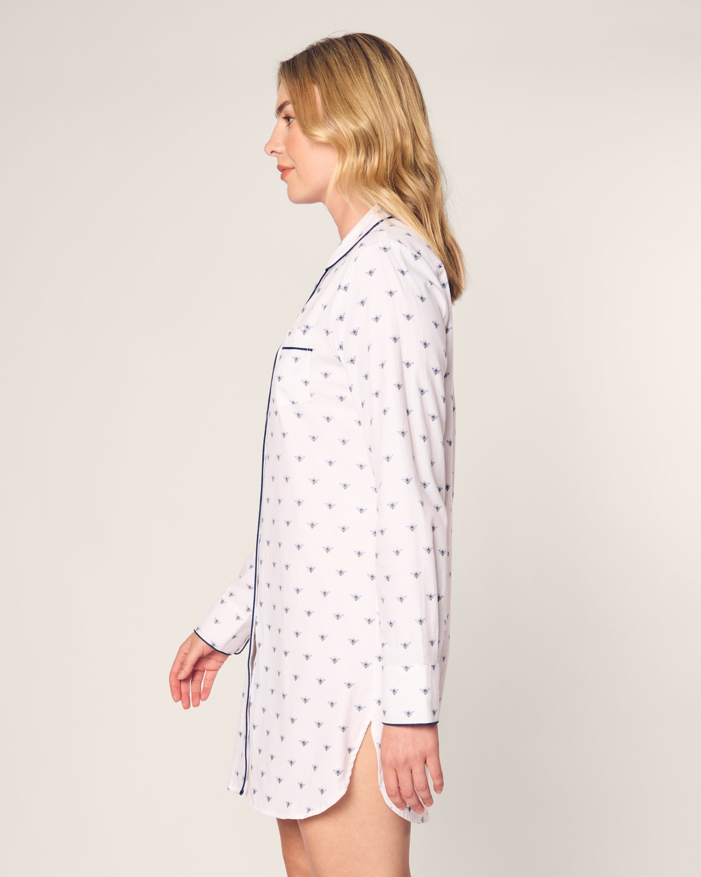 A woman with long blonde hair stands in profile wearing Petite Plumes Womens Twill Nightshirt in Buzzing Bees, featuring dark blue piping. Made from soft cotton twill, it complements the plain light gray background.