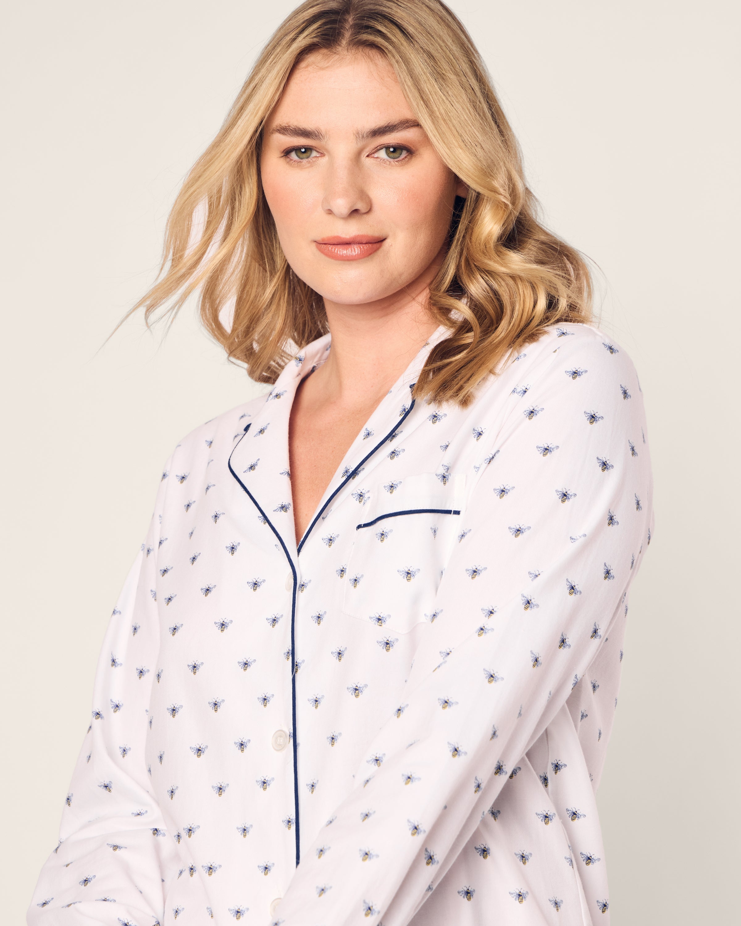 A person with shoulder-length blonde hair is wearing Petite Plumes Womens Twill Nightshirt in Buzzing Bees, a moisture-wicking white pajama top with blue floral patterns and dark piping along the collar and buttons.