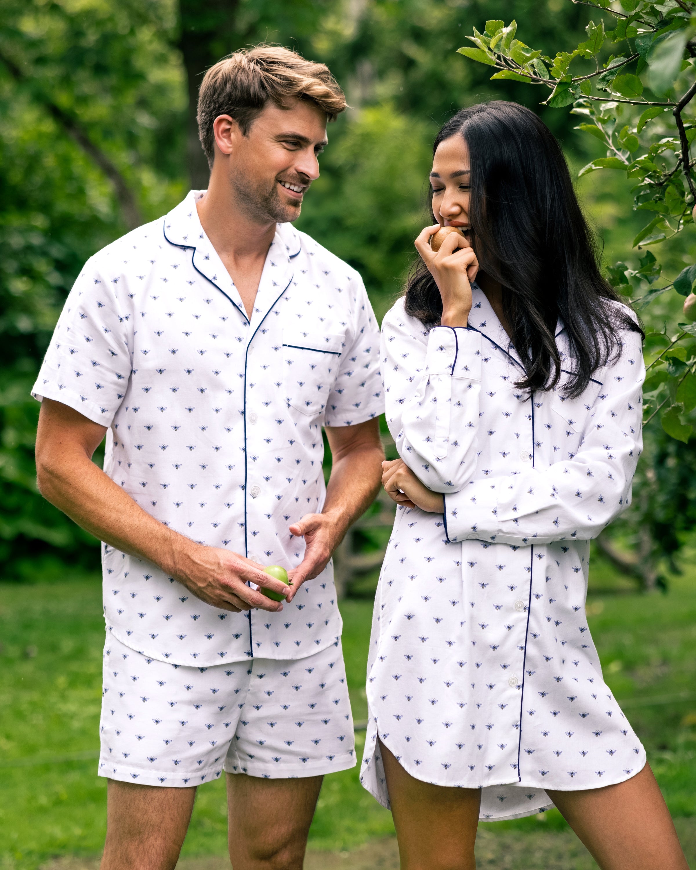 A man and woman in matching white Petite Plume pajamas with the Buzzing Bees print stand on grassy ground, surrounded by lush greenery. The man holds a small fruit while the woman smiles, covering her mouth with her hand.