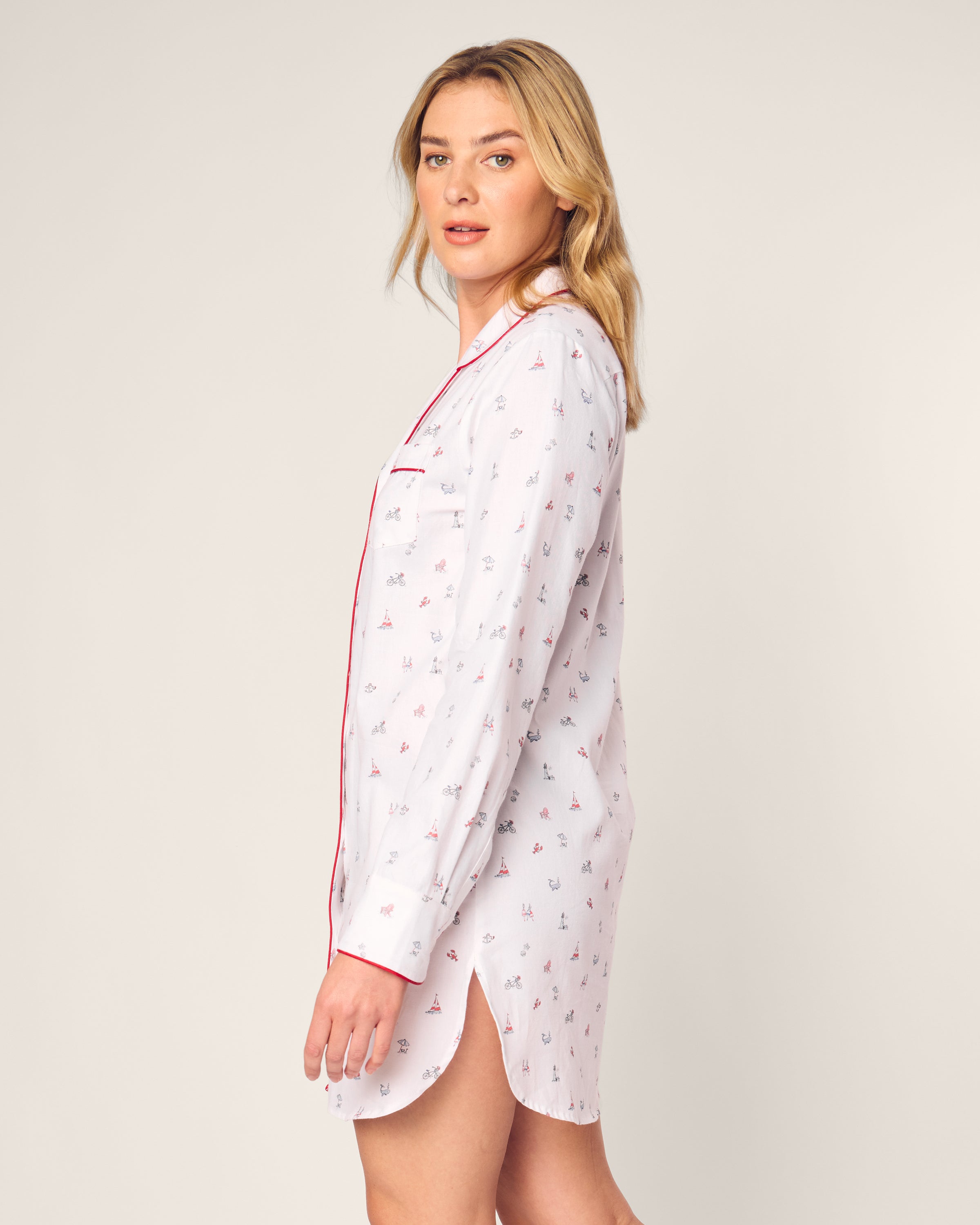 A person with long blonde hair wears Petite Plumes Womens Twill Nightshirt in Coastal Charms, featuring a white, long-sleeved cotton design with colorful prints and red piping. They stand sideways, facing the camera against a light background.