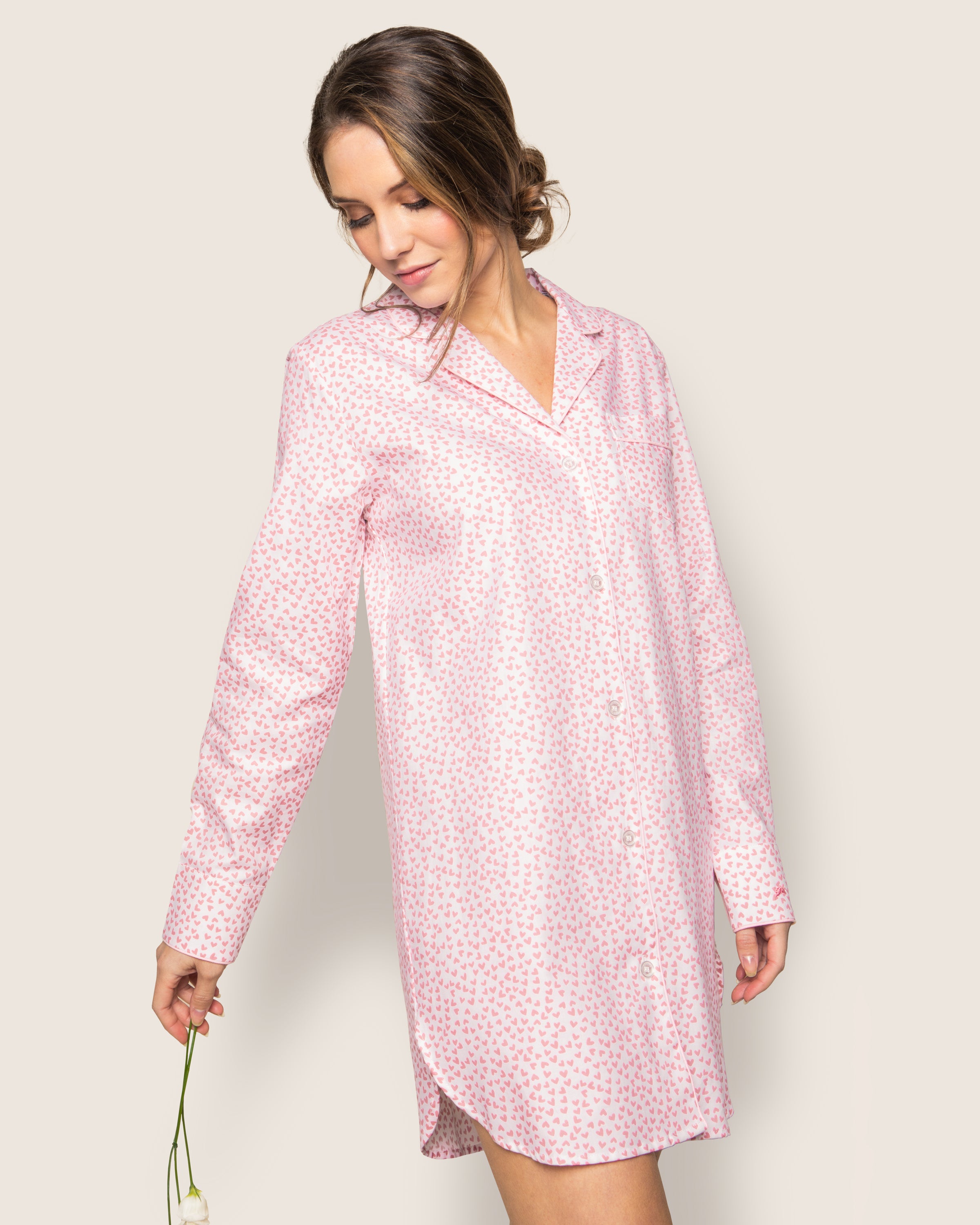 A person in a luxurious Petite Plume Womens Twill Nightshirt in Sweethearts holds a white flower, head slightly down and to the side, with hair tied back against a plain light background.