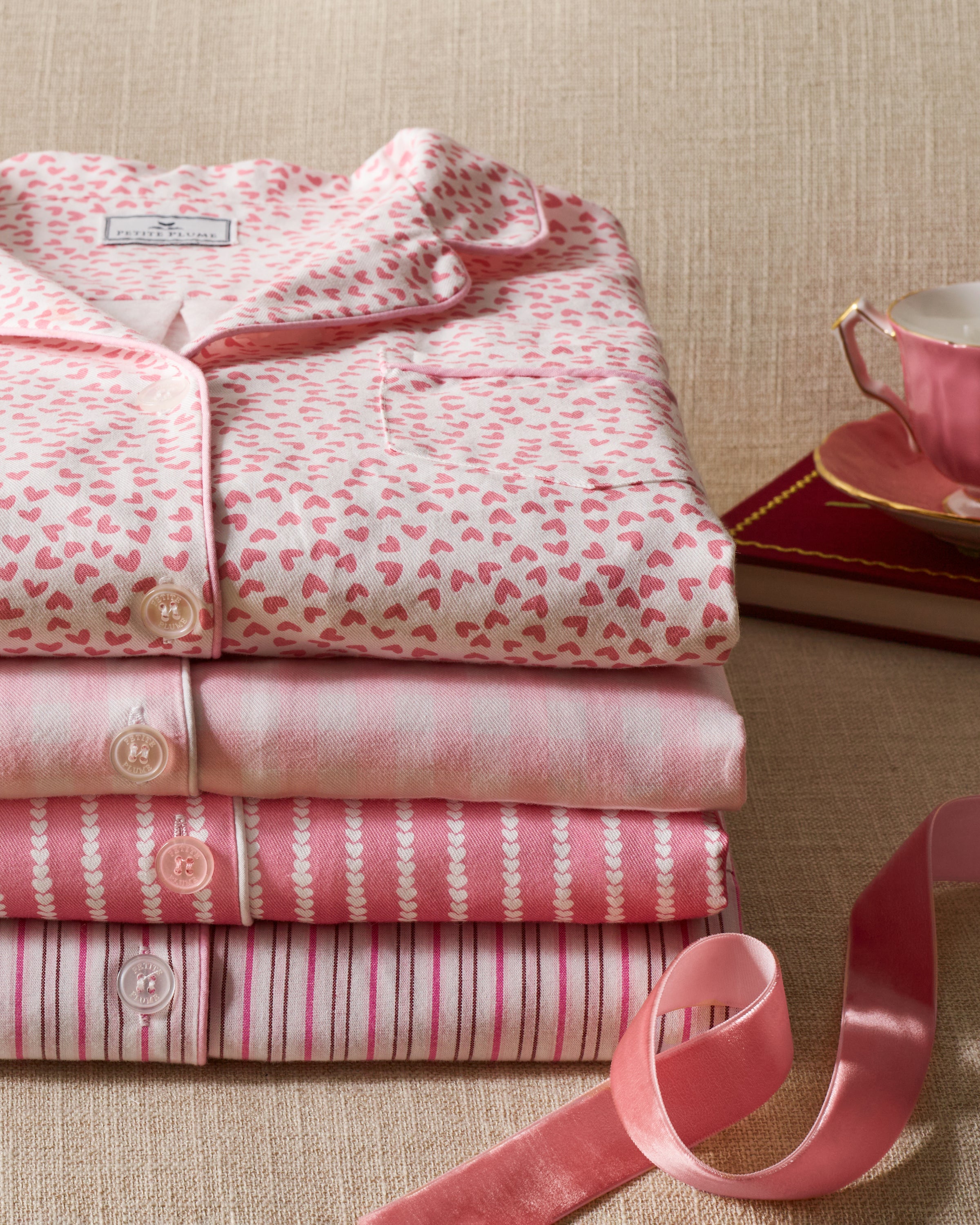 A stack of luxurious Petite Plumes Womens Twill Nightshirt in Sweethearts is neatly folded, alongside a pink teacup and saucer on a book with a pink ribbon, all set against a beige background.