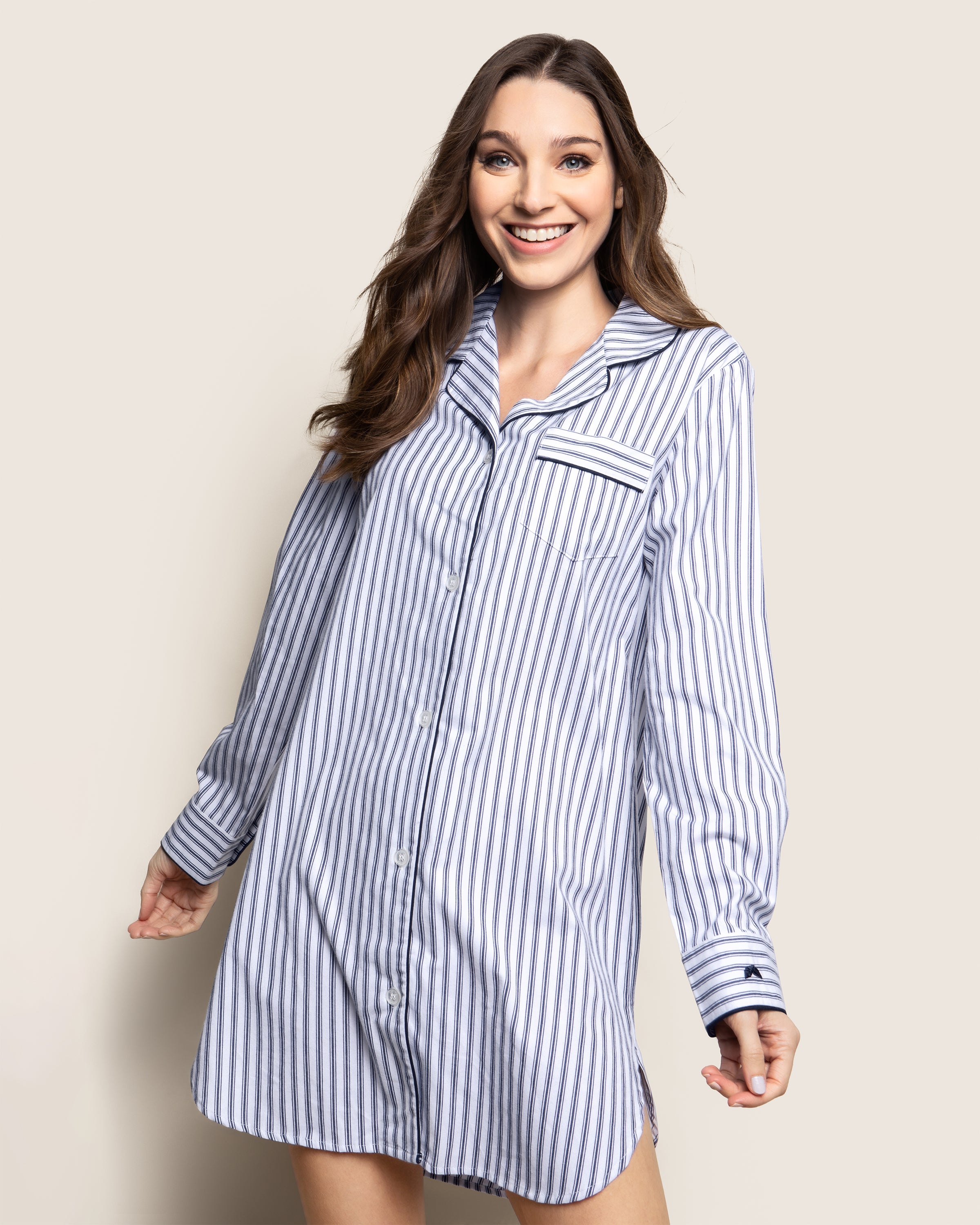 A person wearing Petite Plumes Womens Twill Nightshirt in Navy French Ticking, featuring classic blue and white stripes and a chest pocket, stands smiling with styled-down hair against a plain background, hands casually by the sides.