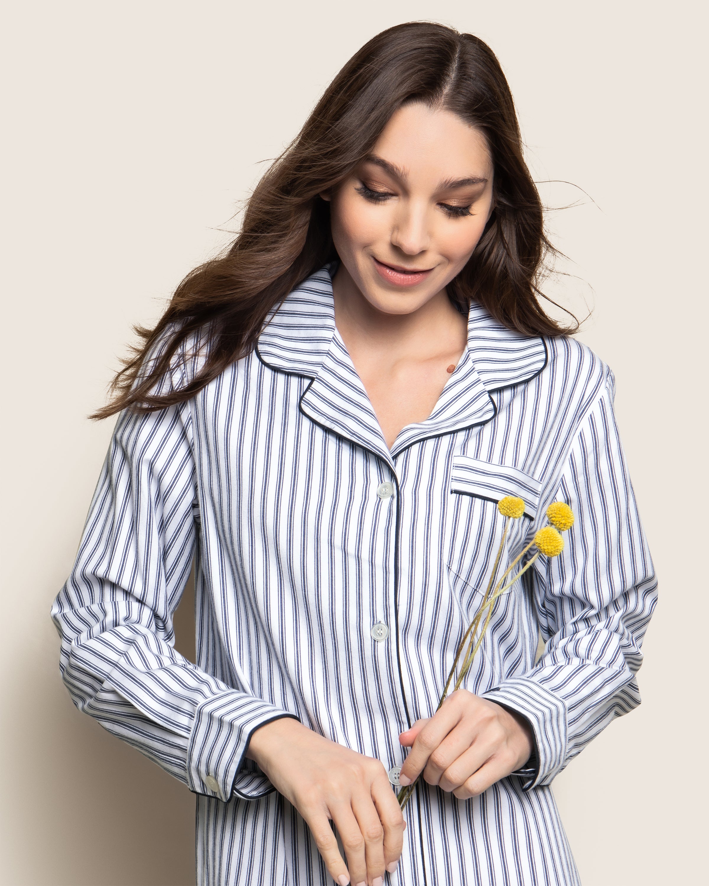 A person with long brown hair wears a Petite Plume Womens Twill Nightshirt in Navy French Ticking. They smile softly while holding small yellow flowers, set against a light background.