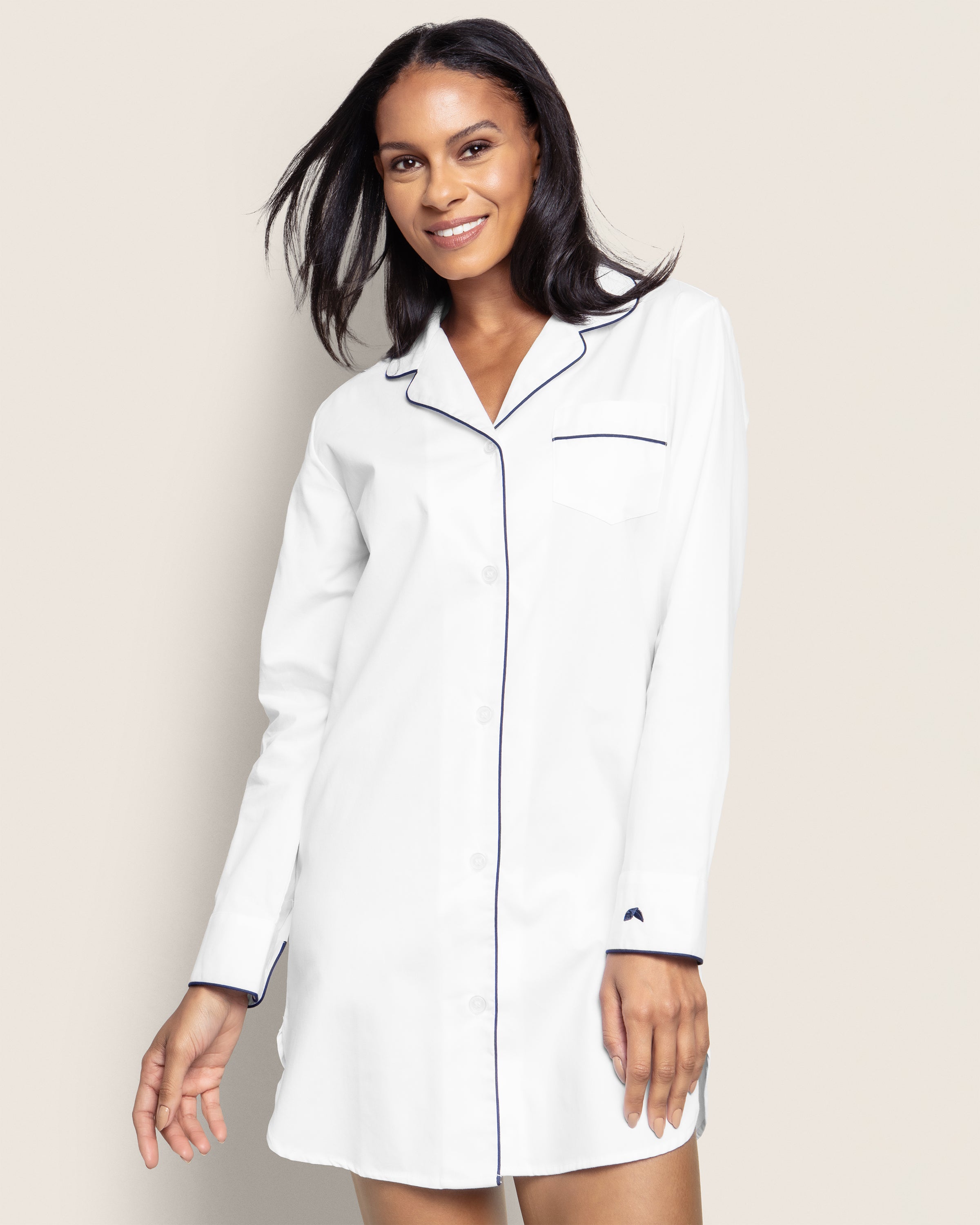 A person smiles in a Petite Plume Womens Twill Nightshirt in white with navy piping, featuring 100% cotton and moisture-wicking fabric, a button-down front, and a chest pocket, set against a plain light background.