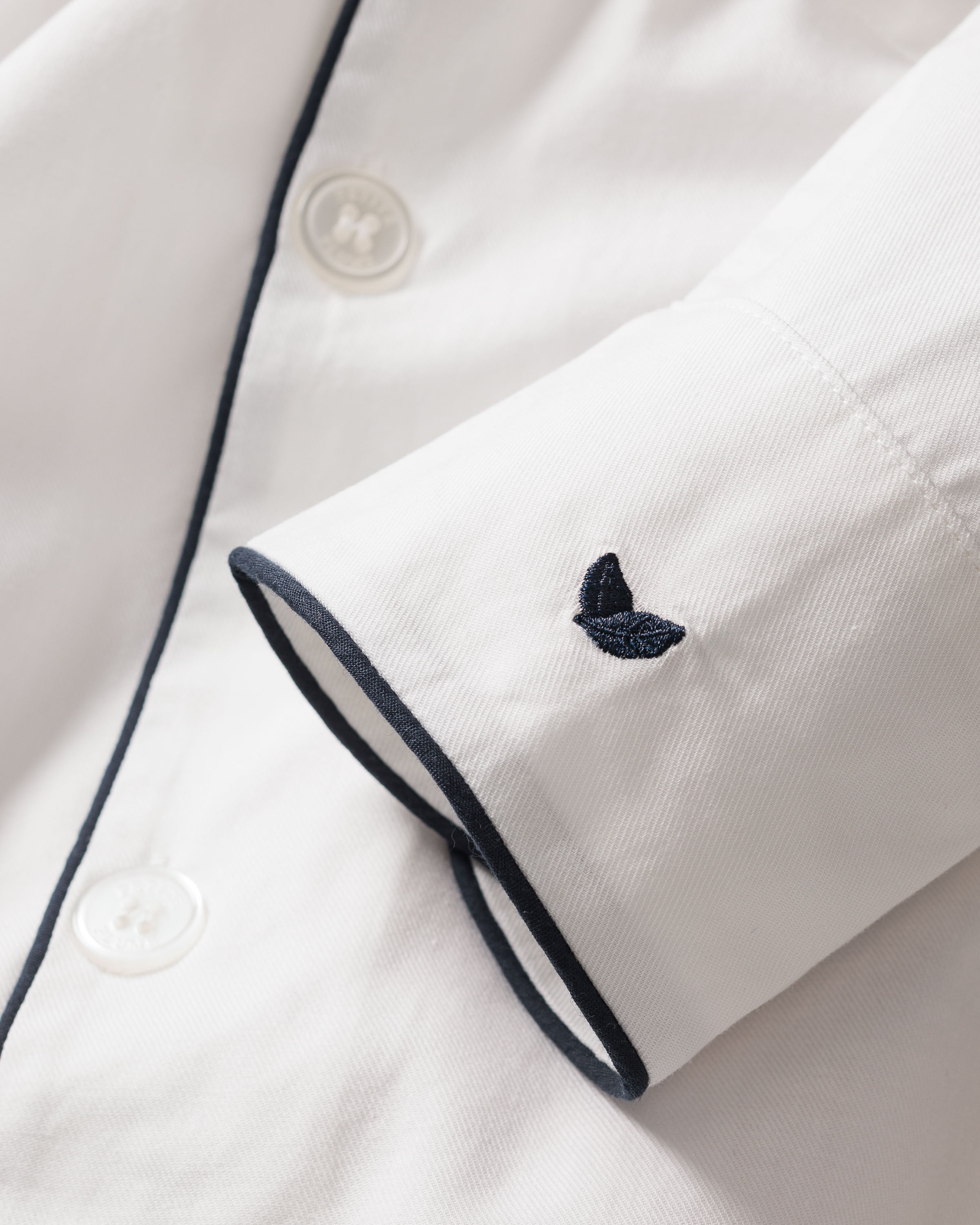 A close-up of the sleeve from Petite Plumes Womens Twill Nightshirt in White with Navy Piping shows 100% cotton twill material, a small navy embroidered butterfly near the cuff, and two white buttons.