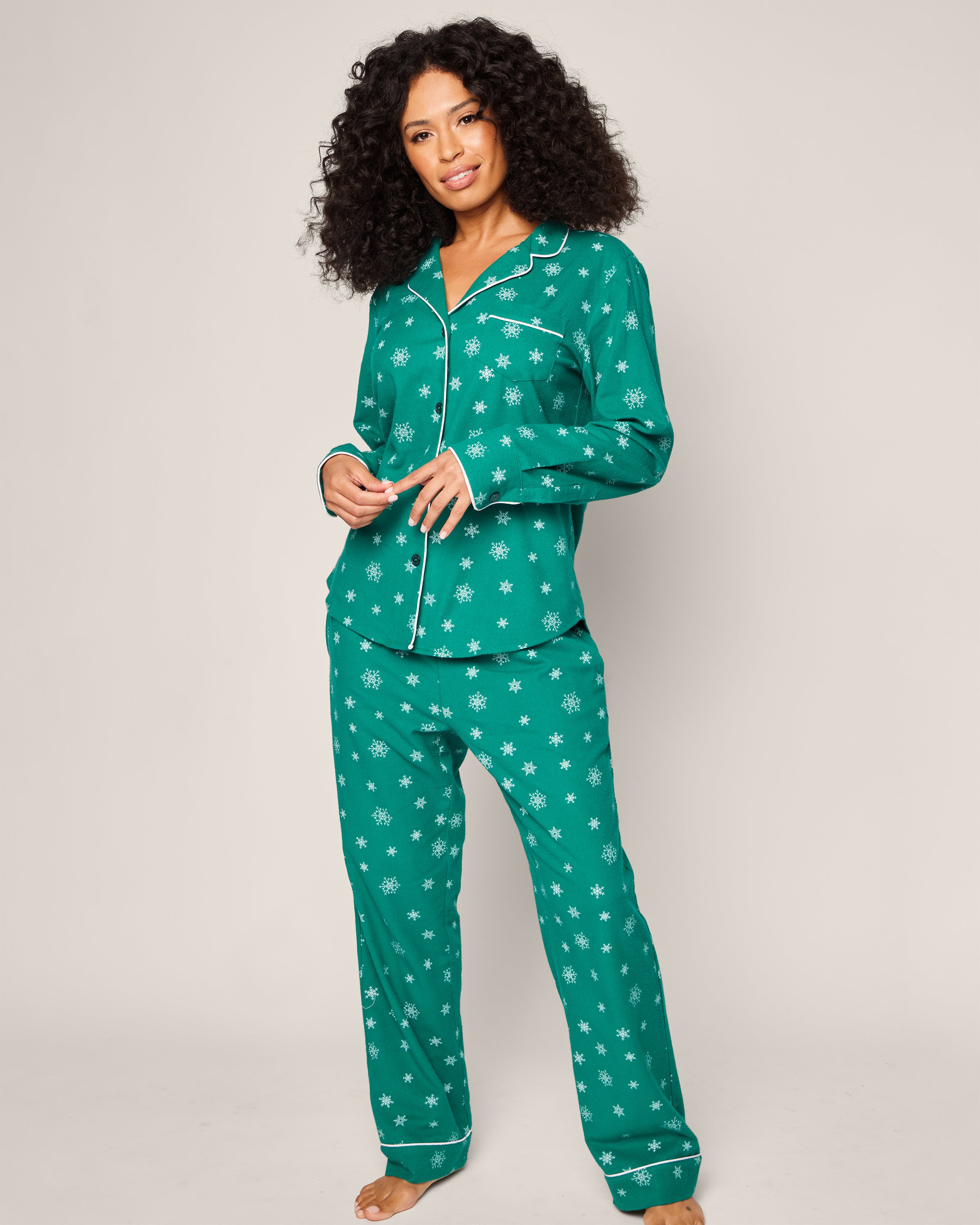 Against a plain background, a woman with long, curly hair and a radiant smile models the Womens Flannel Pajama Set in Emerald Wonderland by Petite Plume. The luxurious cotton sleepwear features an elegant button-up top and matching pants, adorned with white snowflake patterns for ultimate comfort.