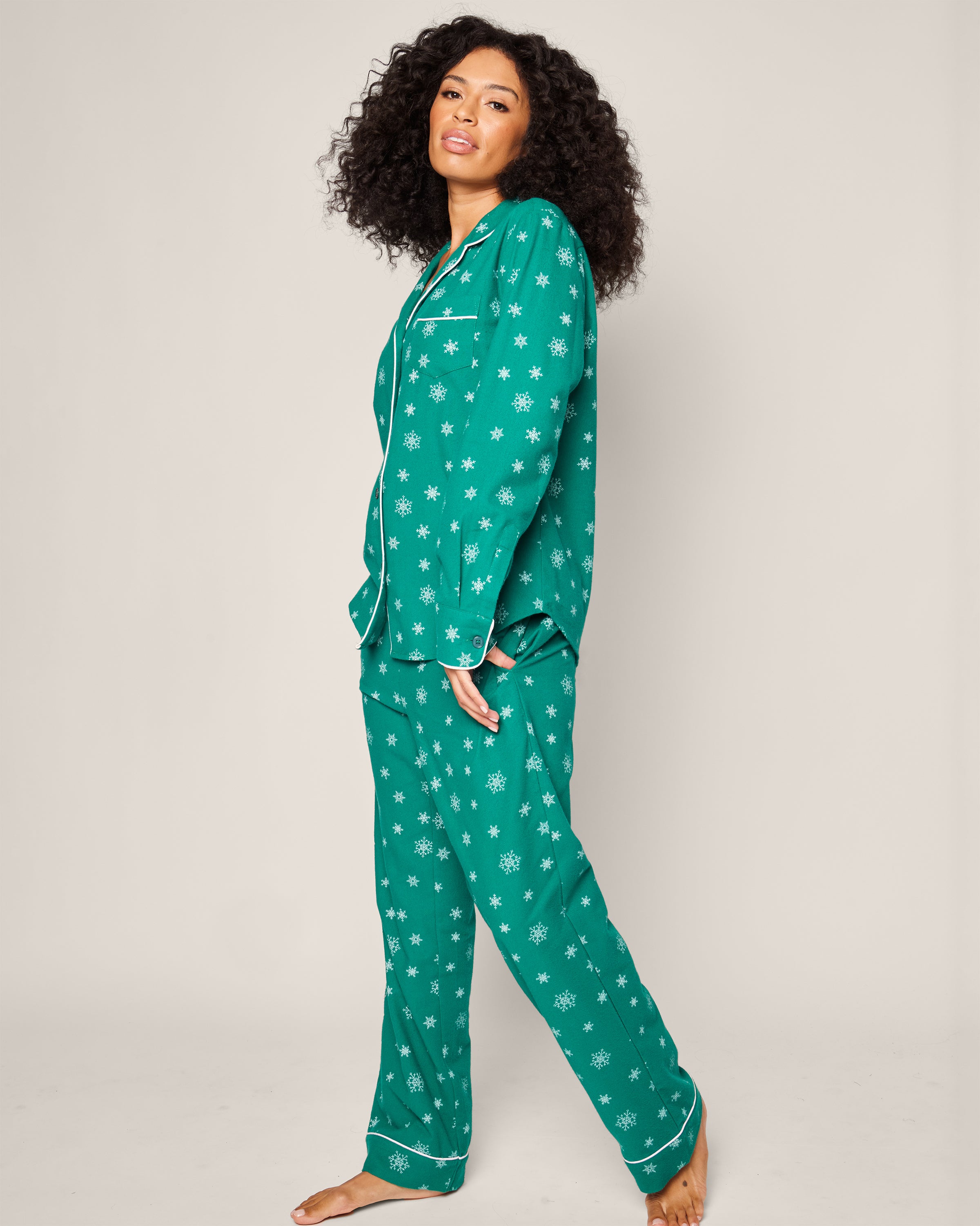 A person with curly hair stands barefoot against a plain light background, smiling slightly while wearing the Petite Plume Womens Flannel Pajama Set in Emerald Wonderland. This luxury cotton sleepwear features an emerald green button-up shirt and matching pants adorned with white star patterns.