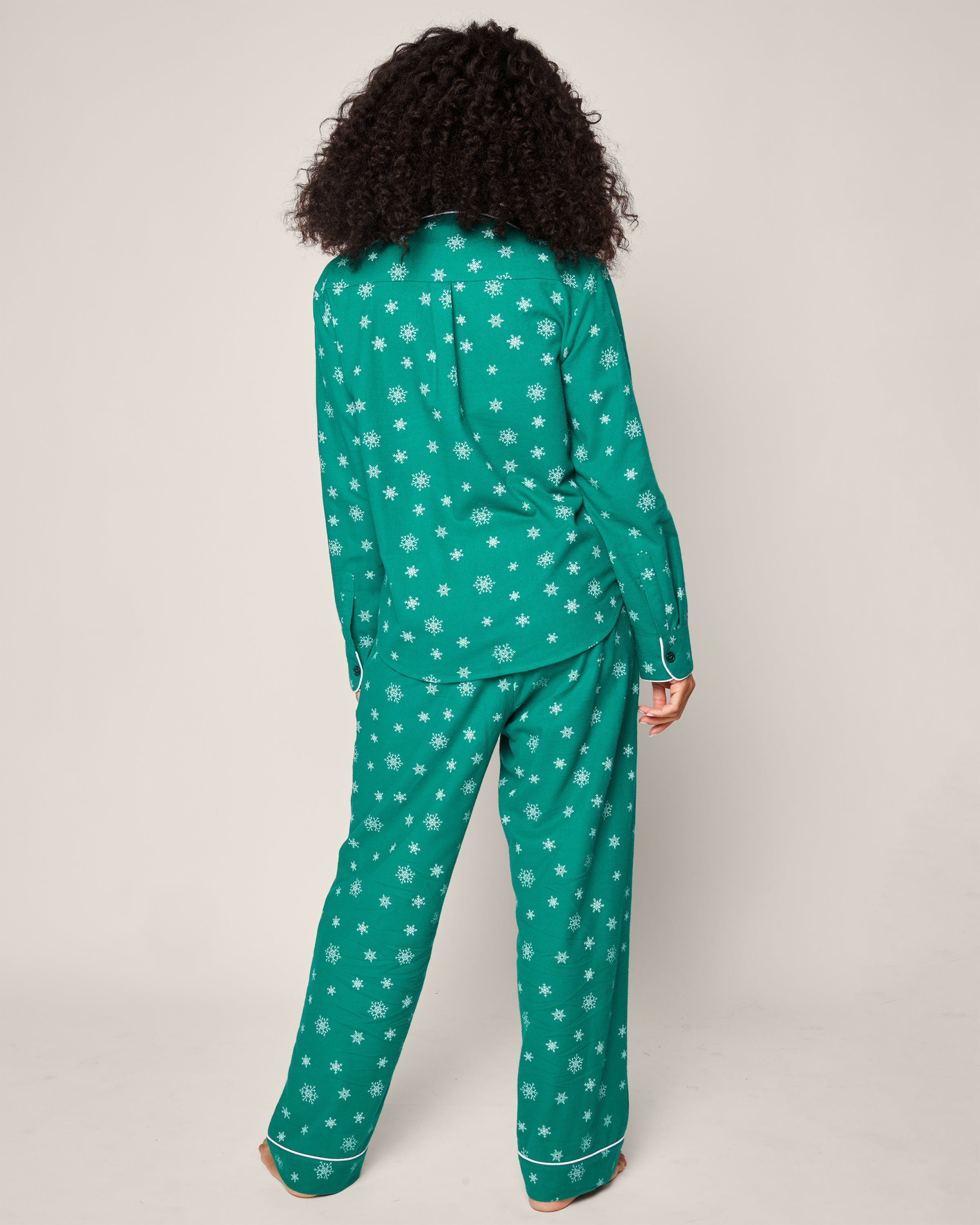 A person with curly hair is turned away, wearing the Womens Flannel Pajama Set in Emerald Wonderland by Petite Plume, featuring white snowflake patterns. The room has a neutral, light backdrop.