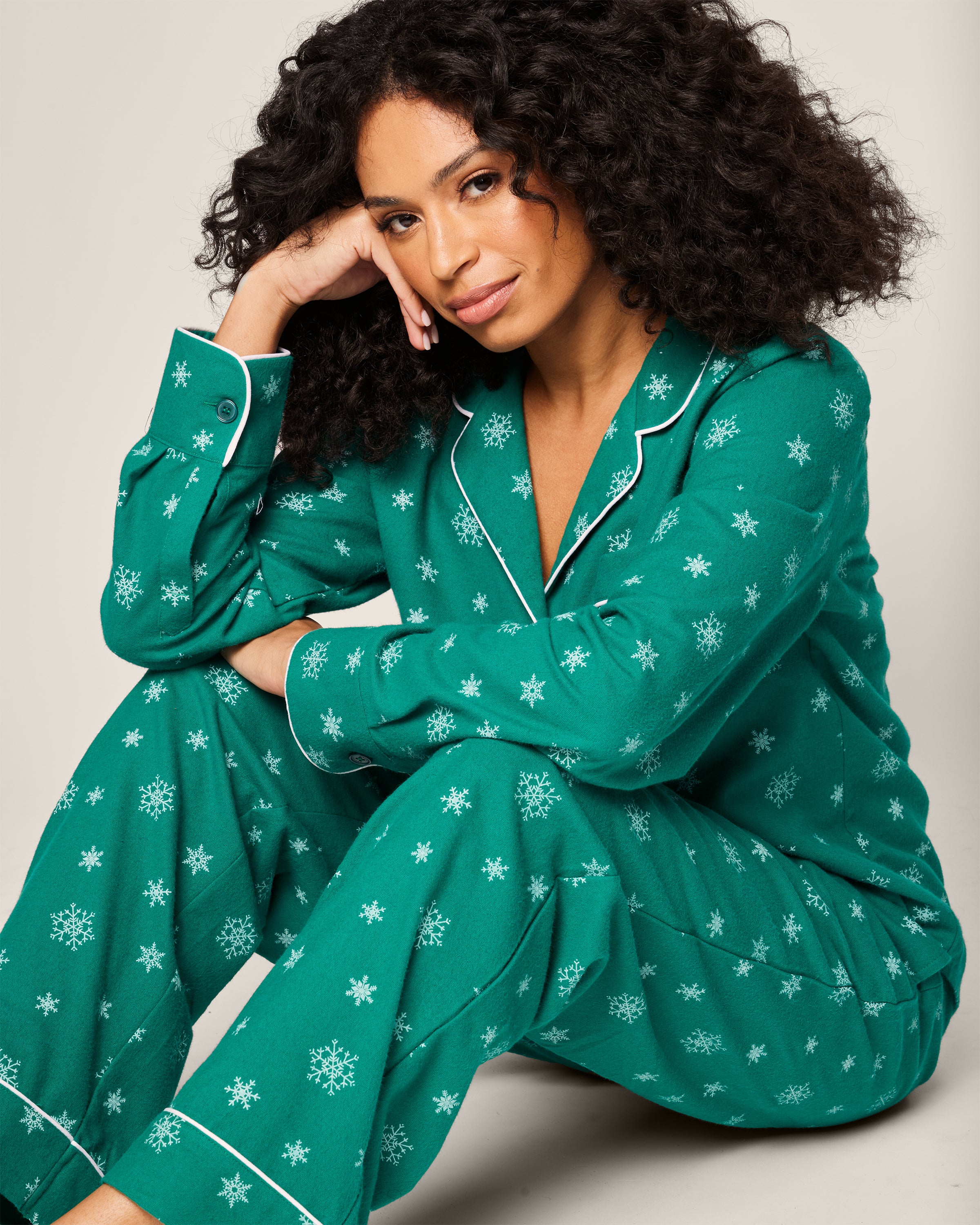 A person with curly hair sits on the floor in Petite Plumes Womens Flannel Pajama Set in Emerald Wonderland, featuring white snowflake patterns and piping. Leaning on one hand, they smile softly against a plain, light-colored background that enhances the cozy luxury of their cotton sleepwear.