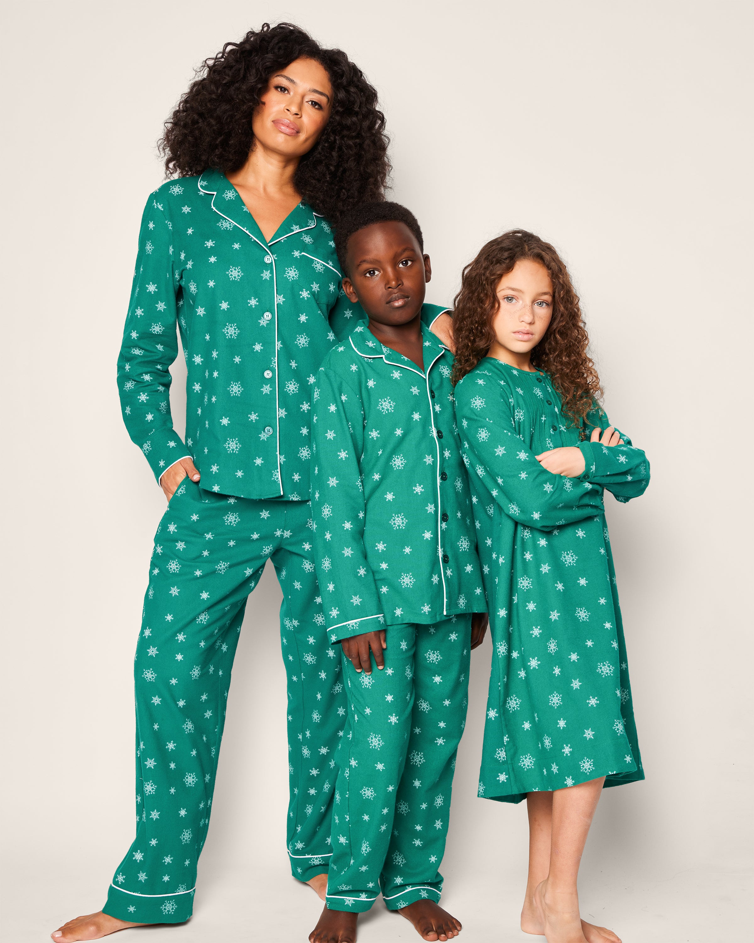 A woman and two children stand together in Petite Plumes Womens Flannel Pajama Set in Emerald Wonderland. The matching sets feature emerald green flannel with white snowflakes, made from the finest quality cotton, as they pose barefoot against a simple backdrop.