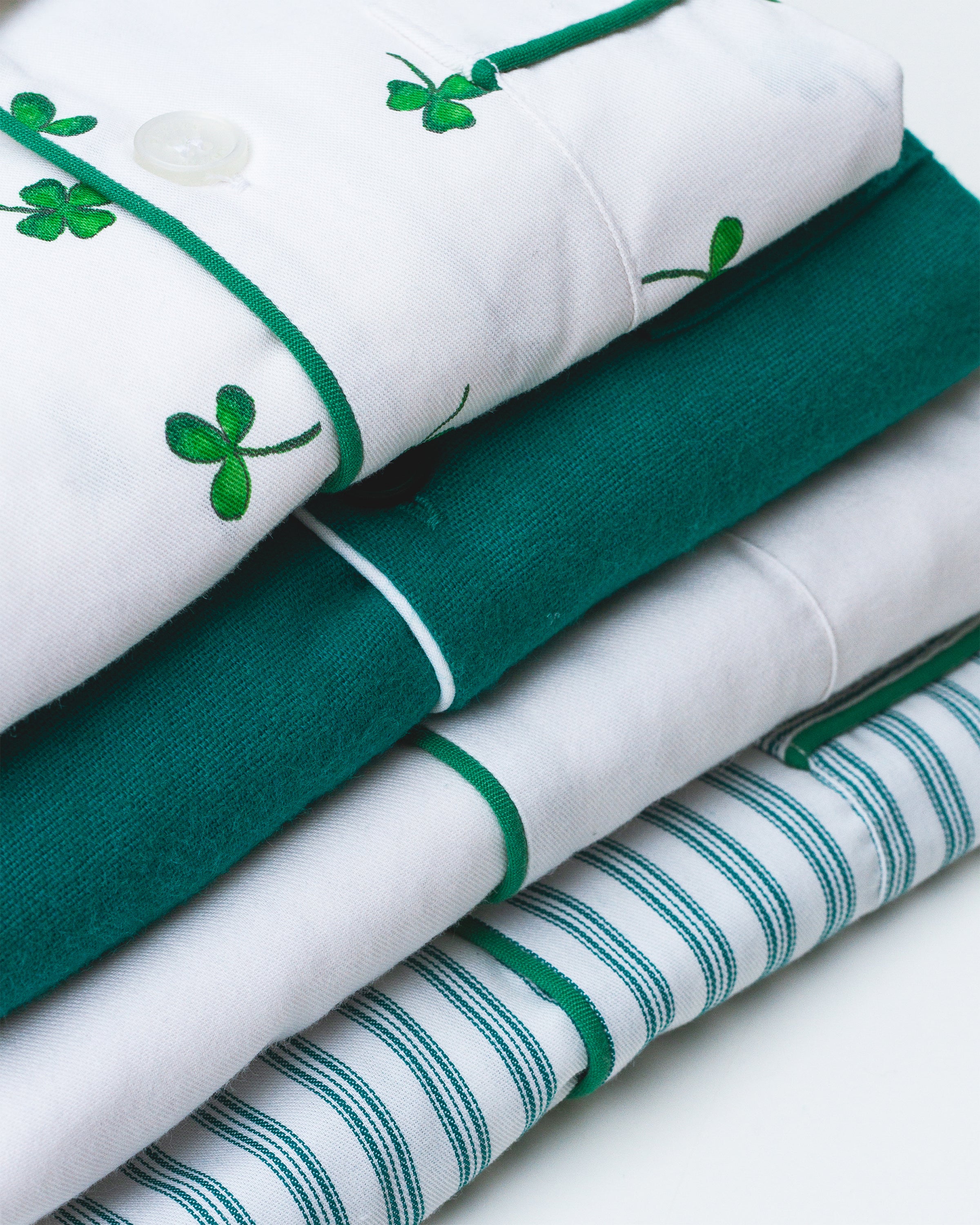 A stack of Petite Plumes Womens Twill Pajama Sets in Emerald Ticking showcases moisture-wicking fabric: the top has green clovers, the second is solid green, the third is white with green trim, and the bottom features bold green and white stripes.